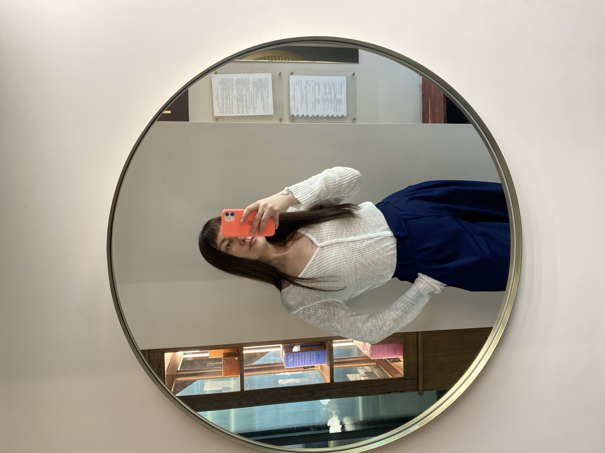 A mirror on a wall shows the reflection of a woman taking a selfie with a pink phone. The woman is wearing a white top and a dark blue skirt. Behind her in the reflection, there is a display case with books and papers on a wall. The location in the city of Votkinsk, Russia, is not determinable from the provided image.
