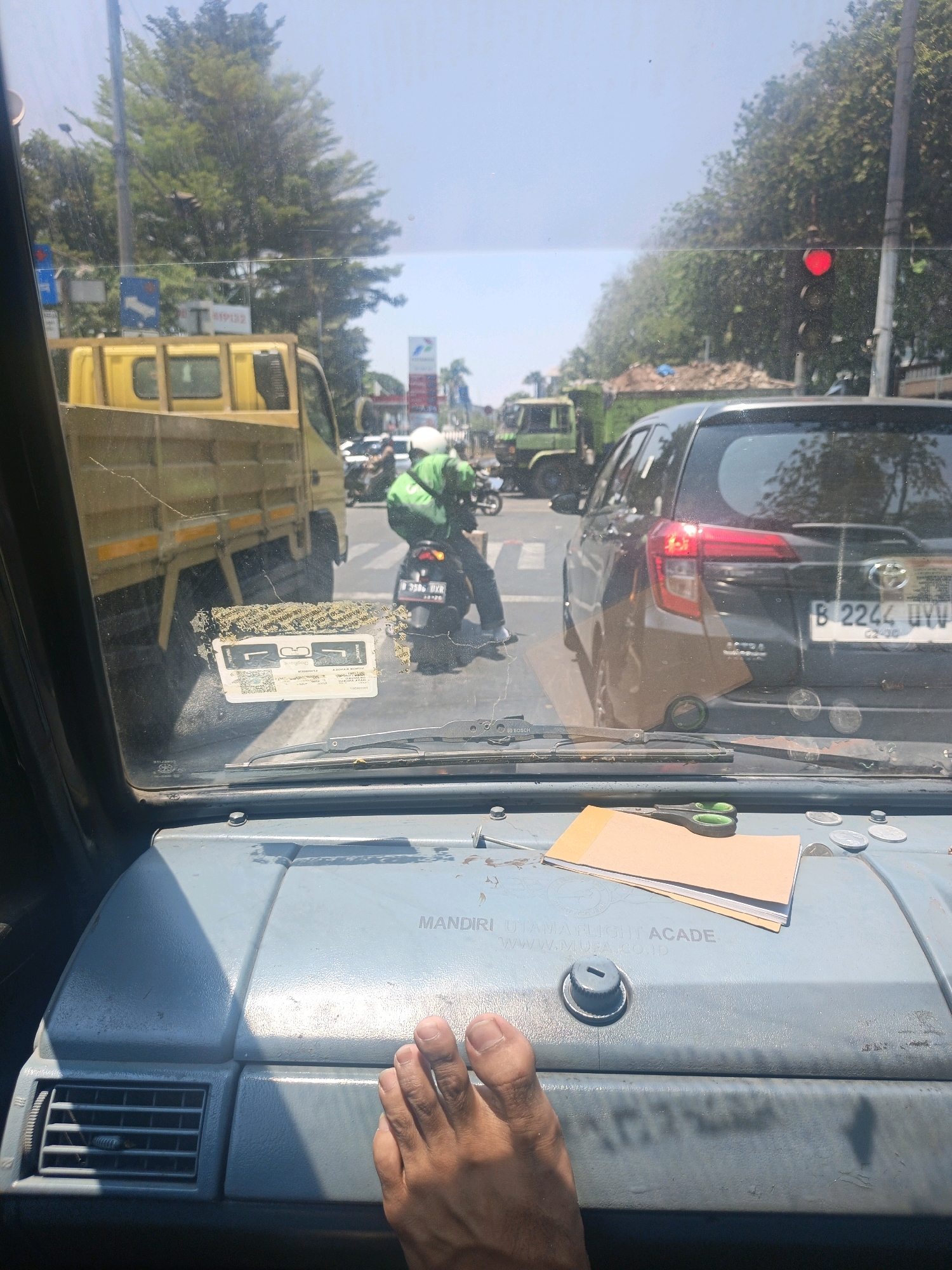 The image is taken from inside a vehicle looking through the windshield. The view shows a street scene in North Jakarta, Indonesia, with moderate traffic. In the foreground, on the dashboard, is a foot, a small stack of papers, a pair of scissors, and several coins. Through the windshield, a yellow truck is visible on the left, and a black car with license plate B 2244 UYY is in front and slightly to the right. A motorcyclist wearing a green jacket and white helmet is to the left of the black car. A traffic light showing red is visible in the background. The environment appears to be a sunny day with trees lining the street.