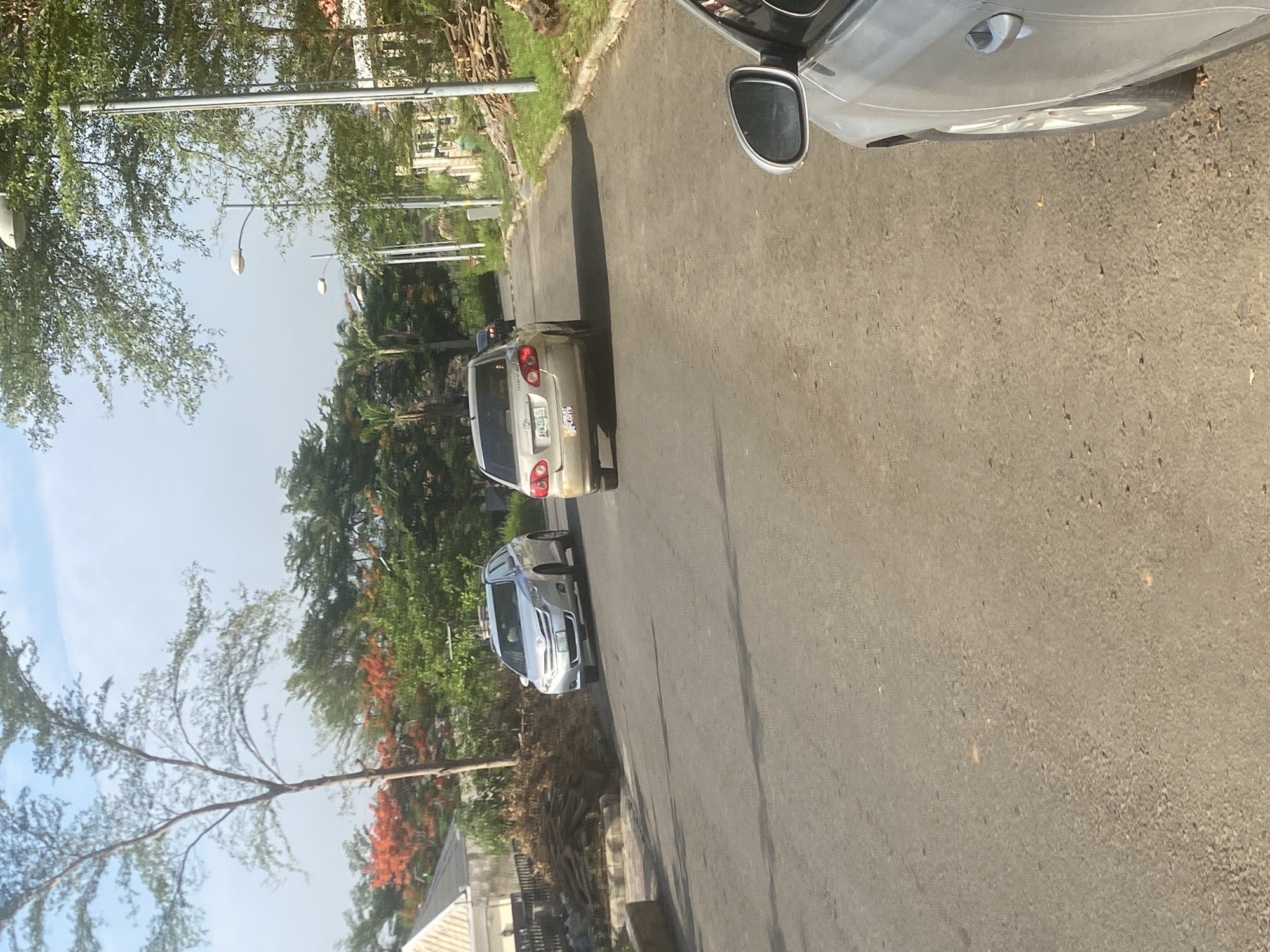 The image shows a street in Unknown, Unknown, with several cars parked on the side. The street is paved with asphalt and there are trees and bushes lining the sides. A grey sedan is parked in the foreground. Two cars, a silver sedan and a silver SUV, are parked behind the sedan. In the background, there is a building or house with a light-colored roof. The weather appears sunny, with a clear sky visible. Street lights are placed on the side of the road.