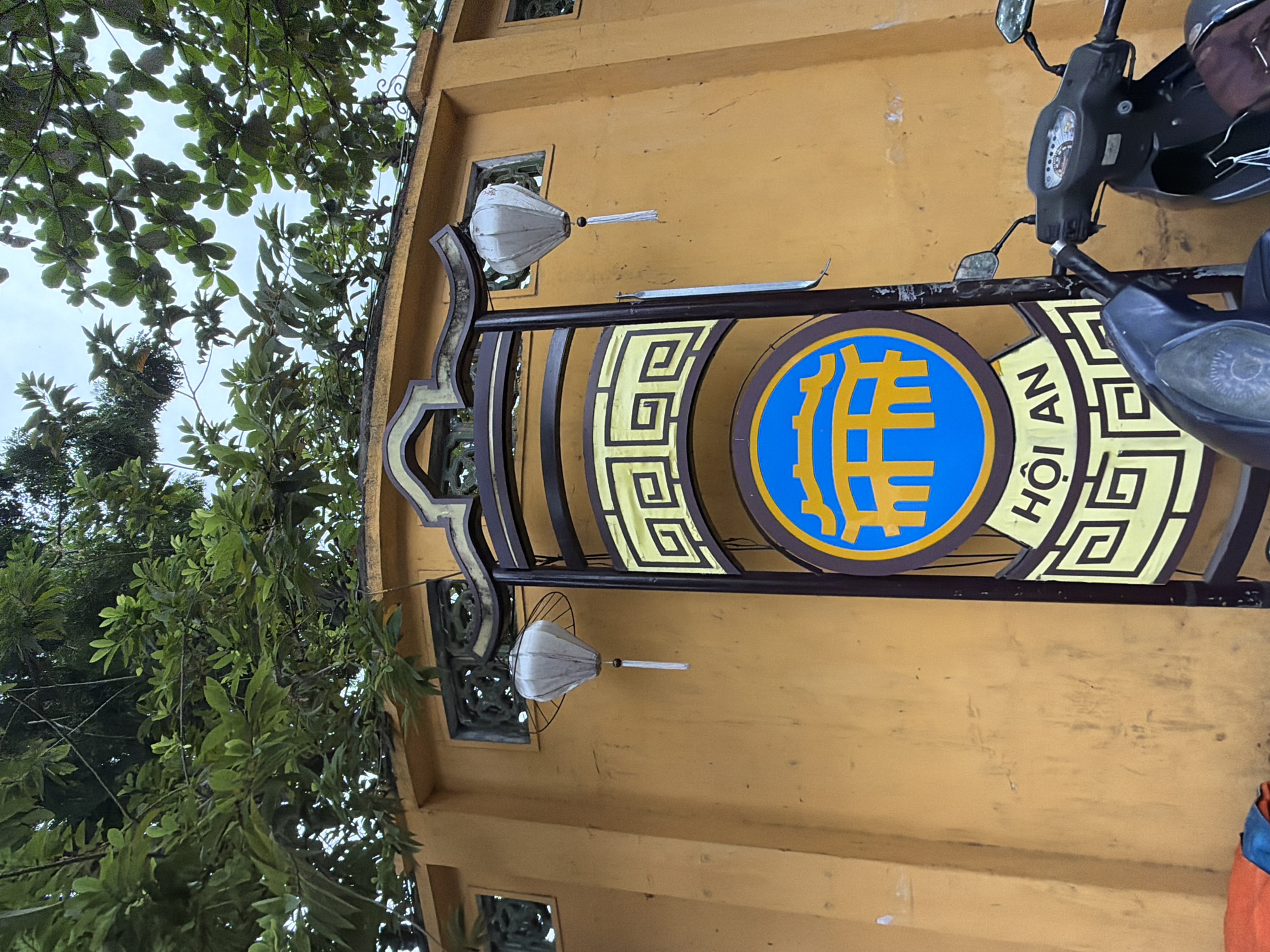 In Hoi An, Vietnam, a traditional sign with a yellow and blue emblem featuring a bridge and the text "HOI AN" stands against an ochre-colored wall. Two white lanterns hang near the top of the wall, and the foliage of trees partially obscures the sky. The lower portion of the image shows parts of two parked motorcycles.