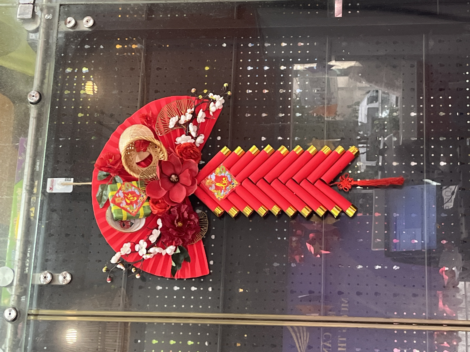A festive arrangement is displayed against a dark, reflective surface dotted with a grid pattern, likely a shop window or display case. The arrangement consists of a red paper fan adorned with red flowers, white blossoms, and a miniature woven basket. A green and yellow object, resembling a cake or wrapped gift, sits on the fan. Attached to the right of the fan is a series of red, tube-like objects with gold ends, arranged to form an arrow shape, finished with a red tassel. The reflection in the dark surface shows elements of a cityscape, suggesting an urban location. The composition indicates the image was taken in Bac Nha Trang Ward, Vietnam.