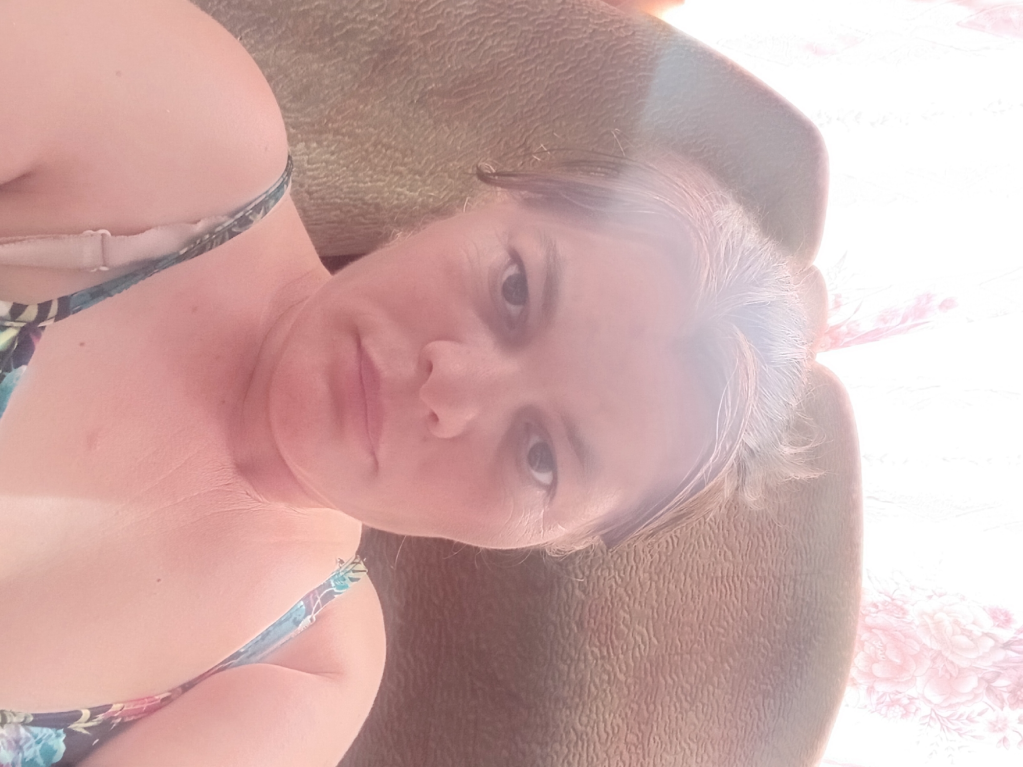 A selfie of a woman is captured in an indoor setting. The woman is positioned in the center of the frame, with her head and shoulders visible. She has light brown hair that appears to be slightly messy, and her face is turned slightly towards the camera. Her eyes are open, and she has a neutral expression. She is wearing a patterned top with thin straps. The background consists of a textured surface, possibly a couch or chair, and a wall with a floral pattern. The lighting suggests it might be daytime, with natural light entering from the side. The overall scene appears casual and intimate, typical of a personal selfie.