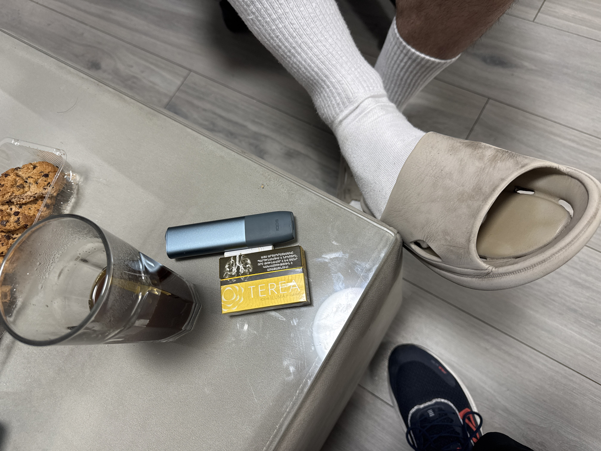 An overhead shot reveals a cluttered scene atop a grey table. A half-empty glass of dark liquid sits next to a plastic container of chocolate chip cookies. An IQOS electronic cigarette and a pack of TEREA cigarettes are also on the table.
A person wearing white socks and beige slide sandals has their foot resting near the table's edge. Another person's blue and white running shoe is visible near the first person's foot.
The scene is indoors, on a grey tiled floor. It cannot be determined from the image if it is in Yerevan, Armenia.