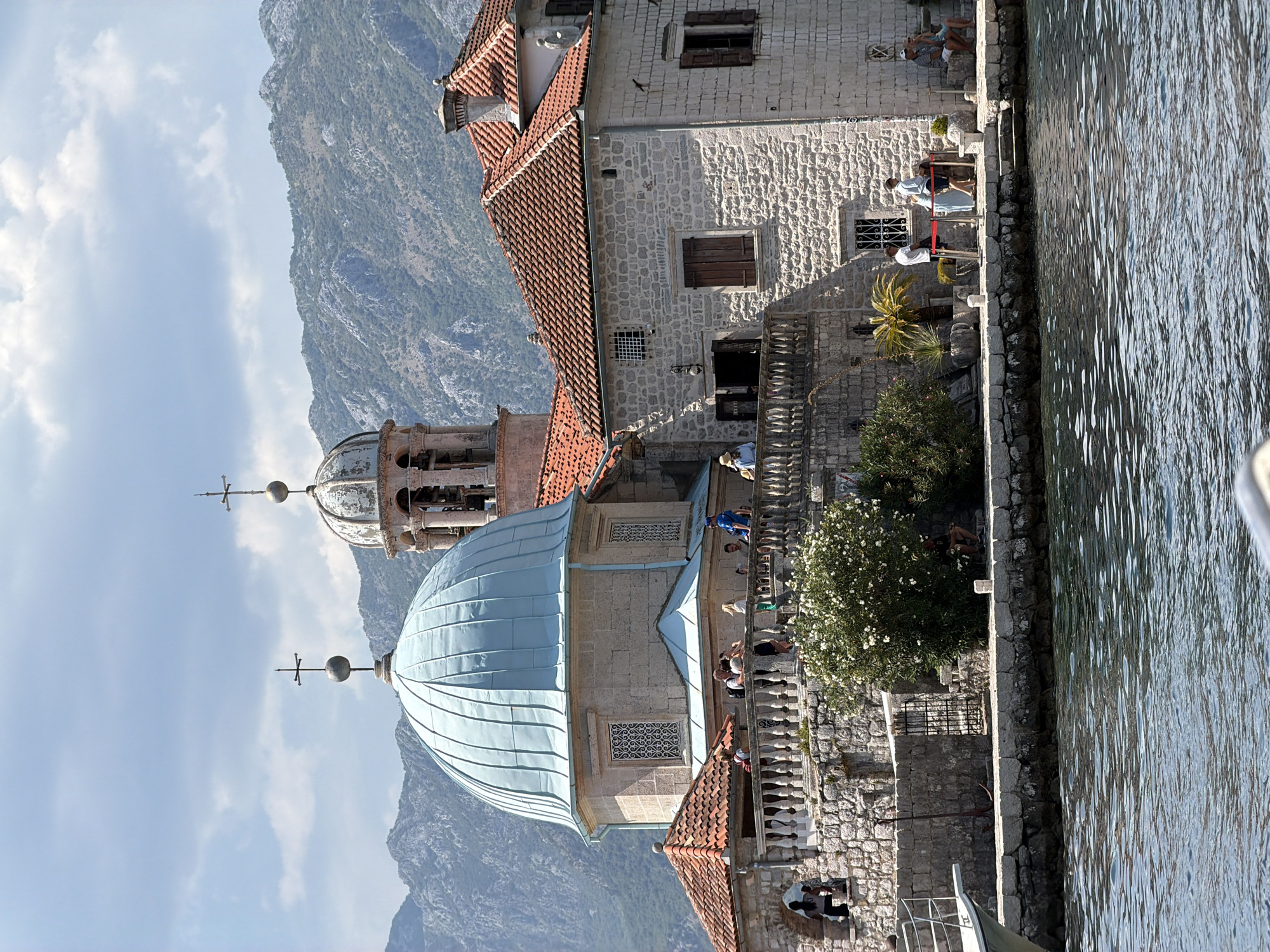 The image depicts the Church of Our Lady of the Rocks, an iconic island church situated in the Bay of Kotor, off the coast of Perast, Montenegro. The church features a distinctive light blue dome and a bell tower, with traditional red-tiled roofs on adjacent buildings. The structures are constructed of stone, with intricate stonework visible on balconies and walls.
Several individuals are present on the church grounds and along the waterfront. On the balcony of the main building, a group of people are seen conversing and observing the surroundings. To the right, on the waterfront promenade, a few individuals are seated, while others stand near a roped-off area. A boat's bow is visible in the lower-left corner, suggesting a maritime perspective.
The background is dominated by a majestic mountain range, its slopes covered in vegetation and rock formations, under a partly cloudy sky. The water in the foreground is calm with gentle ripples, reflecting the light. An anchor is propped against a stone wall on the left side of the image, and a metal gate leads to a set of steps behind it. The overall scene captures a serene and picturesque view of the historic island church.