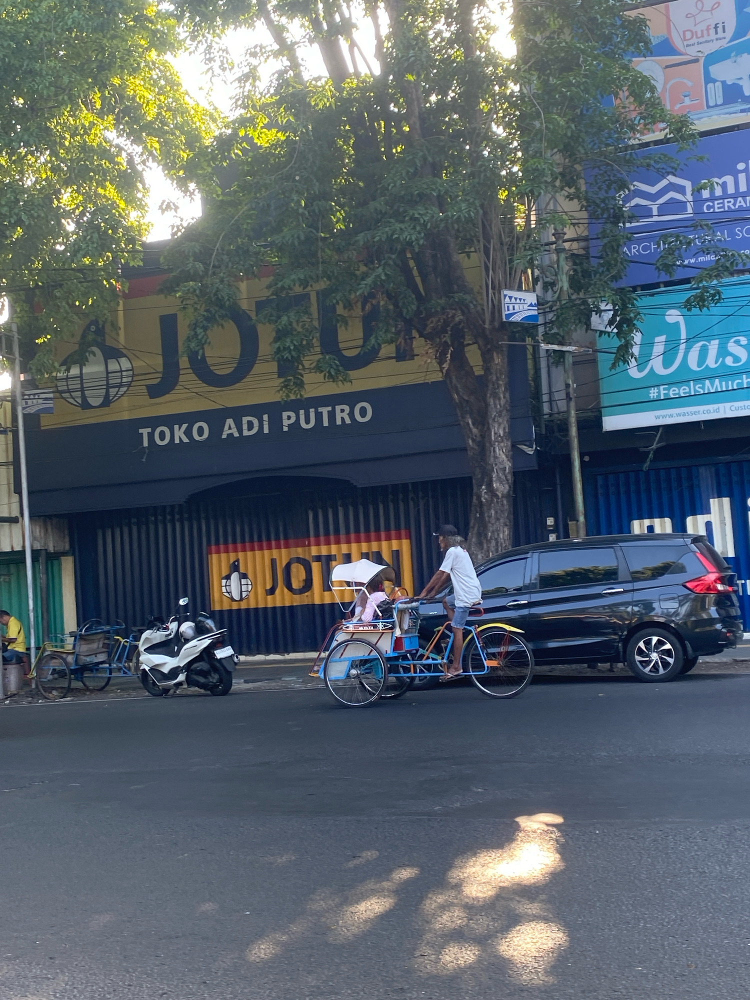 The image depicts a street scene in Kediri City, Indonesia, likely on a sunny day given the strong shadows. A commercial building with a prominent yellow and navy blue facade is visible, featuring the signage "JOTU" and "TOKO ADI PUTRO." In front of the building, a motorized rickshaw, also known as a becak, is in motion. A person is pedaling the becak, which is carrying at least two passengers under a white umbrella. To the left of the becak, a white scooter is parked. Further left, a man in a yellow shirt is seated on a stool next to a wheeled cart. A black multi-purpose vehicle is parked on the right side of the street, alongside the becak. The environment includes trees with lush green foliage casting dappled sunlight on the road. Several other commercial signs are partially visible on adjacent buildings, including one with the word "Wasser" and "#FeelsMuch". Power lines are strung overhead. The road surface appears to be asphalt.
