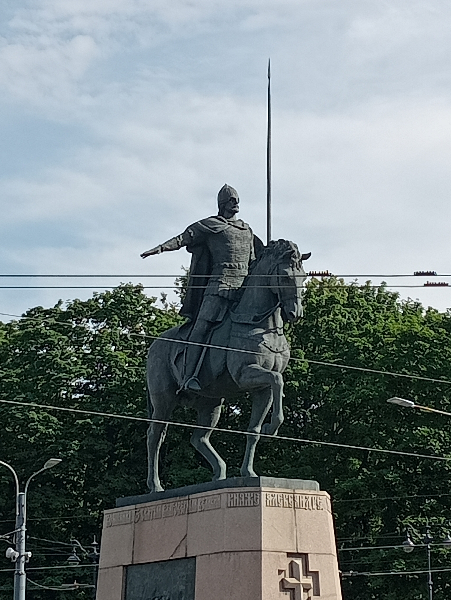 A tall equestrian statue of a man in armor holding a spear stands against a bright, partly cloudy sky. The statue is dark gray and appears to be made of bronze or a similar material. The man is depicted wearing a helmet and a cape over his armor, with his left arm extended outwards and his right hand on the reins of the horse. The horse is also dark gray and shown in mid-stride.
The statue is mounted on a large, multi-tiered stone pedestal. The pedestal has some inscriptions and a carved symbol on its front, which resembles a cross with an additional horizontal bar.
In the background, lush green trees form a dense foliage that partially obscures the sky. Several thin, dark wires are strung horizontally across the image, suggesting the presence of power lines or telephone lines. A street lamp with a modern design is visible on the left side of the image, and another street lamp is visible on the right side, further indicating an urban or suburban setting.
The lighting suggests it is daytime, likely midday given the brightness of the sky. The weather appears to be fair, with some clouds present. The overall scene is static, with no discernible activity other than the representation of movement in the statue itself. The location is not explicitly identified but appears to be an outdoor public space.