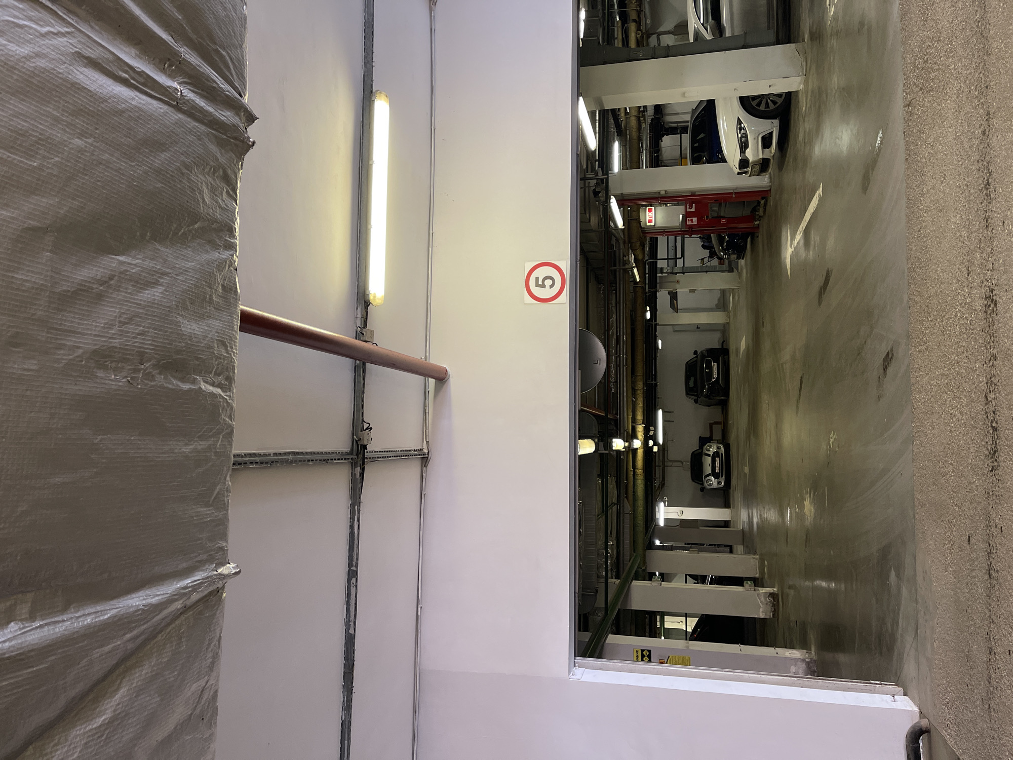 The image depicts an indoor parking garage, likely in Moscow, Russia given the presence of Cyrillic text in the image and general style. The garage appears to be multi-level, with vehicles parked using hydraulic lifts. There are multiple cars visible in the garage. The walls are white and have various fixtures attached, including fluorescent lights. A red sign with the number "5" in a white circle is visible on the wall. The left side of the image shows a structure covered in a gray tarp or sheeting. Overall, the environment is utilitarian.