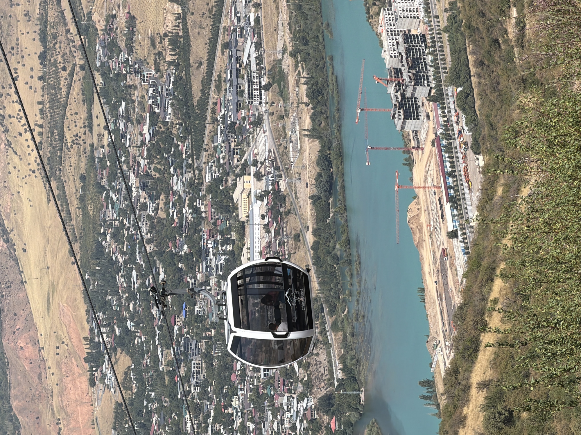 An aerial, eye-level, medium shot shows a cable car cabin moving left to right across the frame, with the city of Charvak, Uzbekistan visible in the background. The cable car is white with black trim and has large glass windows. Inside the cabin, two people are visible. One is a man wearing a light-colored shirt, and the other is a woman wearing a pink top. The cable car is suspended from cables that stretch across the image. Below the cable car, a wide, turquoise river runs horizontally across the frame. Along the riverbank, there is a construction site with several large orange cranes and partially built buildings. Beyond the river, the city of Charvak spreads across a hillside, with numerous houses and buildings, many of which have red or gray roofs. Trees are scattered throughout the city and on the surrounding hillsides. The hills in the far background are dry and barren, with a light brown hue. The sky is clear and bright.