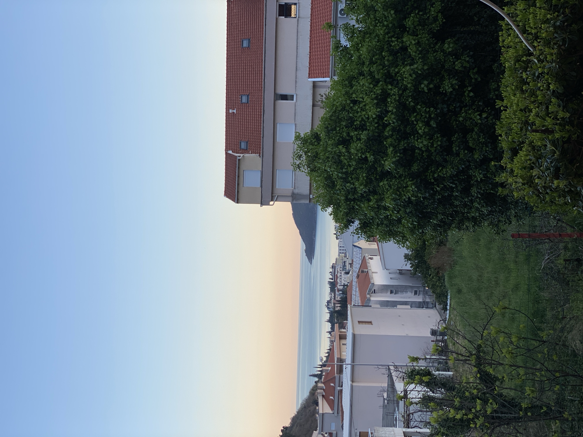 The image depicts a partial view of Budva, Montenegro, at dusk. The sky transitions from blue to a faint orange near the horizon, where the Adriatic Sea stretches out, bounded by a distant mountain. In the middle ground, buildings with red tile roofs intermingle with trees and grass. A multi-story building with beige siding and a red tile roof dominates the upper-left portion of the frame, partially obscured by a large green tree. Fences and vegetation obscure the lower right portion of the image. There is no visible activity in the scene, and the buildings and landscape appear still and silent.