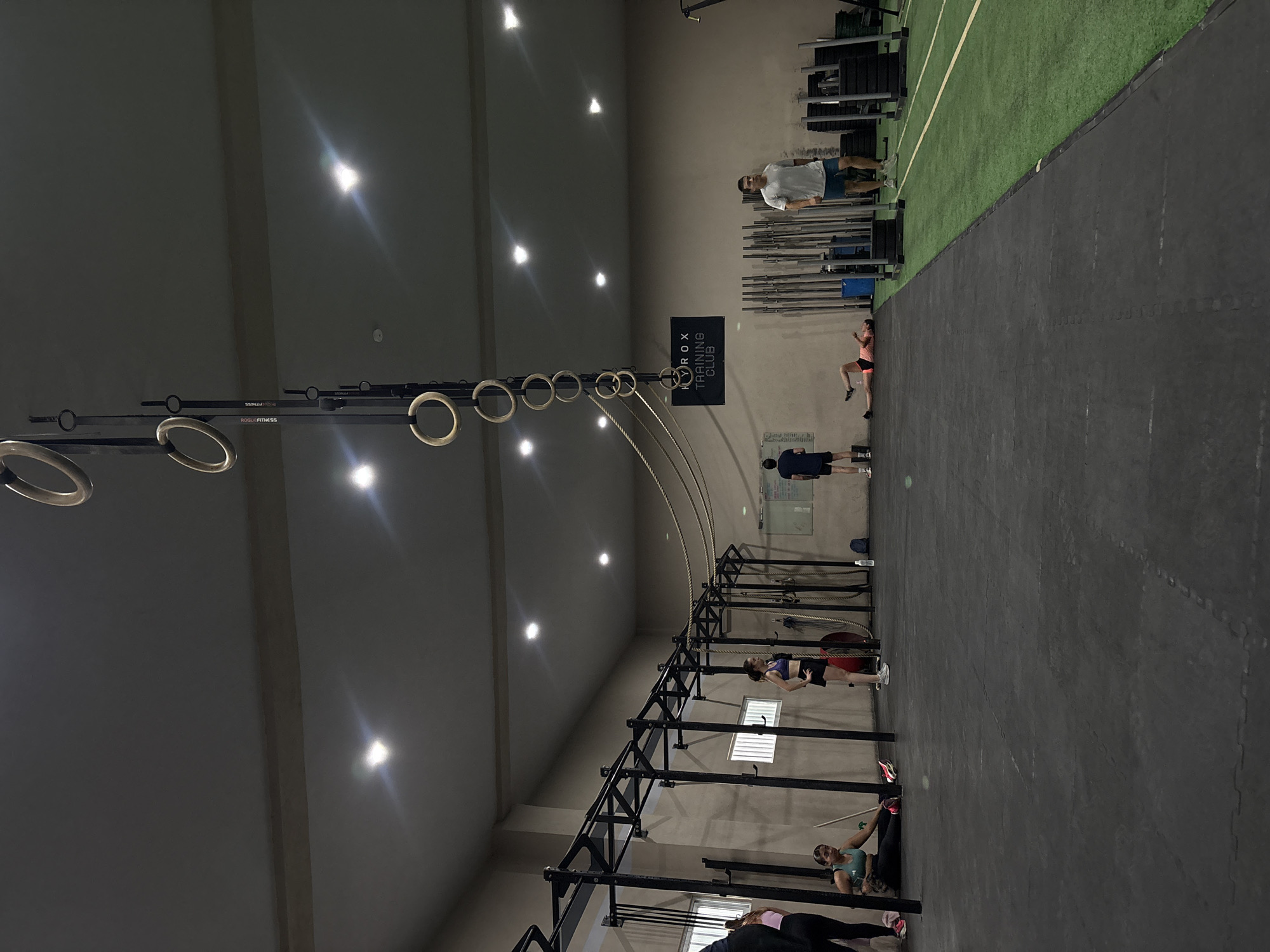 A brightly lit indoor gym in San Bernardino Tlaxcalancingo, Mexico features black rubber flooring and exercise equipment. People are positioned throughout the space, engaged in various fitness activities. Overhead, suspended gymnastic rings and a rope extend across the gym. A section of green turf is visible on the right side. A sign indicates "HYROX TRAINING CLUB".