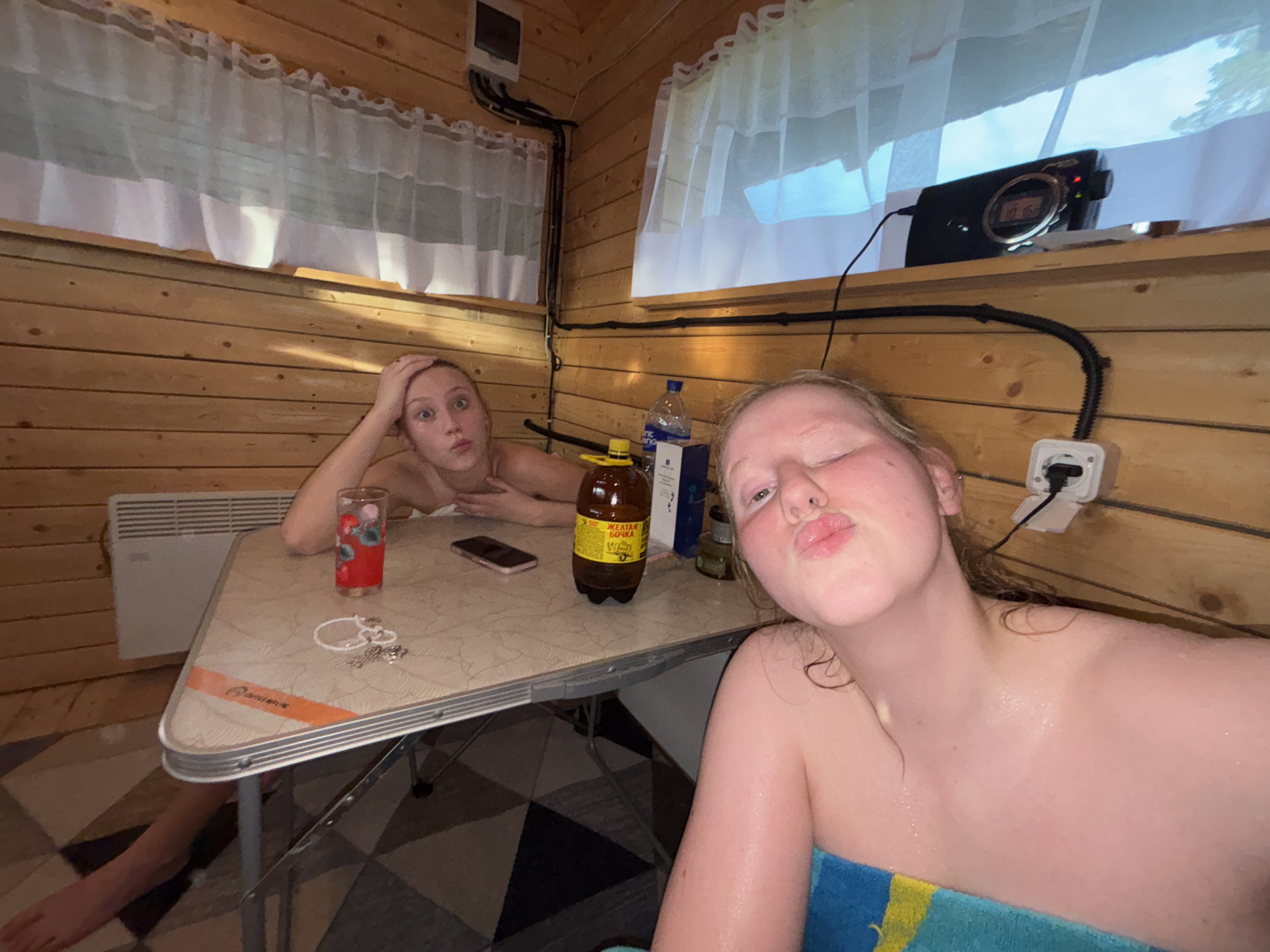 Two young women are posing for a selfie in a wooden room, possibly a sauna or a rustic cabin. The room is lined with wooden planks. The woman in the foreground is closer to the camera, her face close to the lens, with her eyes closed and lips pursed in a kiss. She is wrapped in a blue and yellow towel. The woman in the background is sitting at a small table, holding her head with one hand and looking at the camera with wide eyes. She has a glass of red liquid, a bottle of brown liquid, and a smartphone on the table in front of her. A white electrical outlet is visible on the wooden wall behind the woman in the foreground. A digital clock is mounted on the wall above a window covered by white curtains. The lighting suggests it is daytime, and there are no obvious indications of the weather outside. The overall mood seems playful and relaxed. The label on the brown bottle appears to be in Russian and says "ЖЕЛТАЯ РЕКА" which translates to "Yellow River".