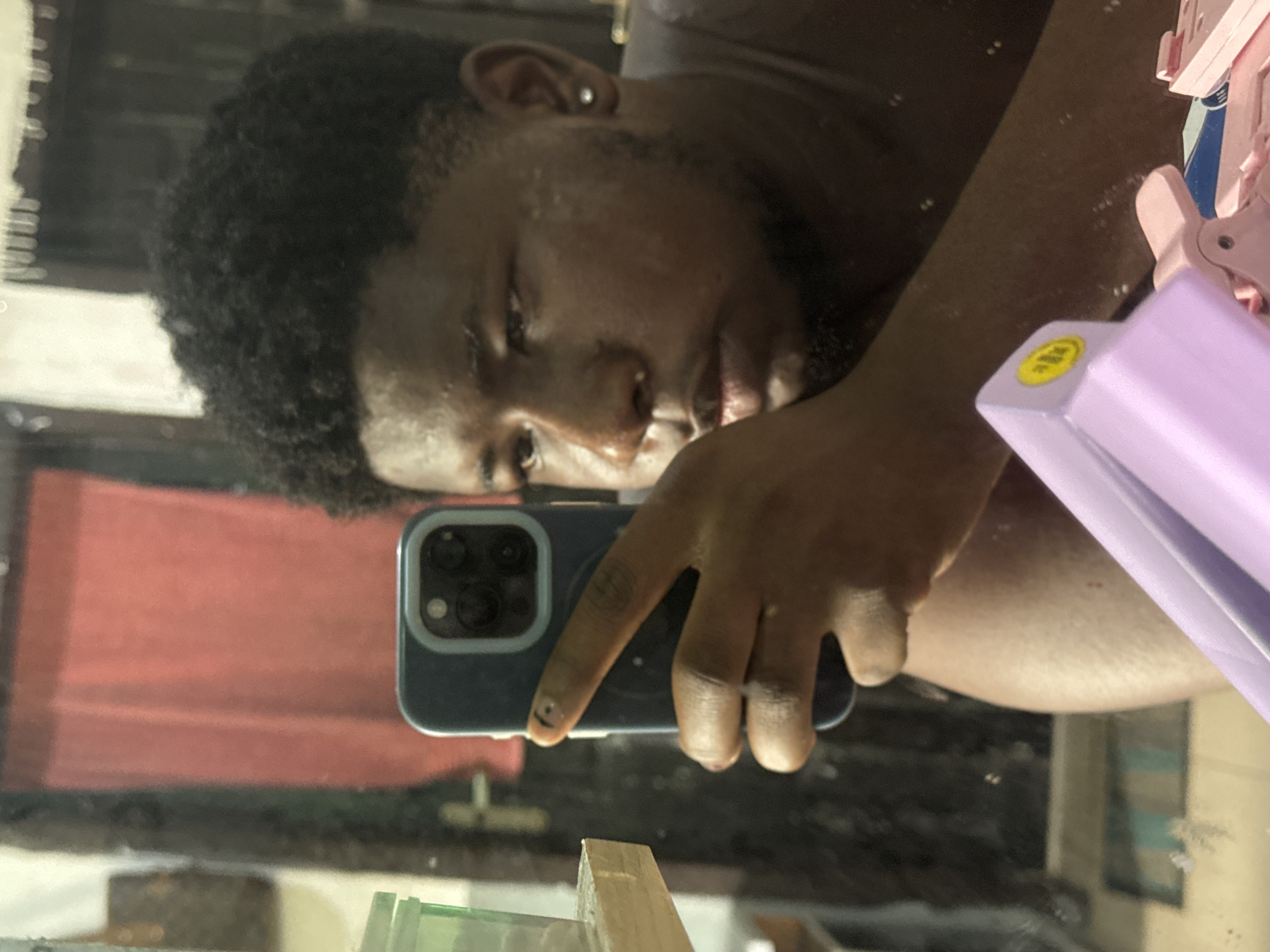 A person with dark skin and curly hair is taking a selfie in a mirror. The person is holding a dark blue iPhone with a triple-lens camera. The reflection in the mirror shows a red wall. There is a lavender-colored object with a yellow sticker in the upper right corner of the image. The location is Asipa, Nigeria.