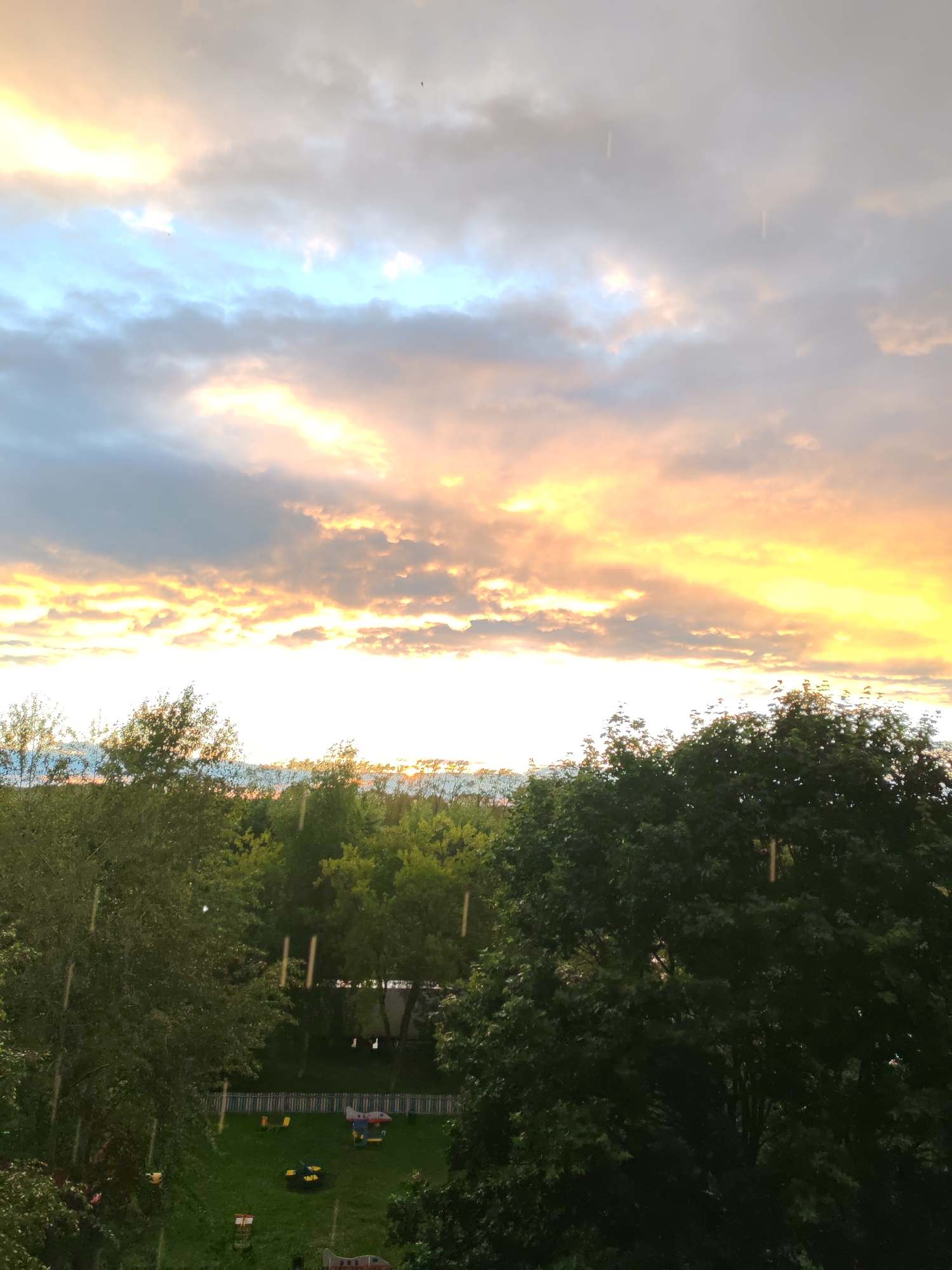 This media file captures a sunset with a cloudy sky, painted in hues of orange, yellow, and blue, with some streaks of white and gray clouds. The sun is setting, casting a warm glow across the sky, suggesting it is late evening. The scene transitions to a lush green landscape filled with trees, creating a natural setting. In the foreground, dense foliage of trees dominates the view, partially obscuring the background.
Beneath the trees, a grassy area is visible, featuring a playground with colorful elements like a red and blue airplane-shaped structure, yellow chairs, and a yellow and black toy. A white picket fence surrounds part of the area, and hints of other play equipment are discernible. The presence of a playground suggests a residential or park area.
The weather appears to be somewhat unsettled, with visible streaks that could indicate falling rain or perhaps a visual artifact in the image. The overall mood is serene and tranquil, amplified by the soft lighting of the sunset. The location is identified as Sokol, Russia. There is no visible text in the media file.