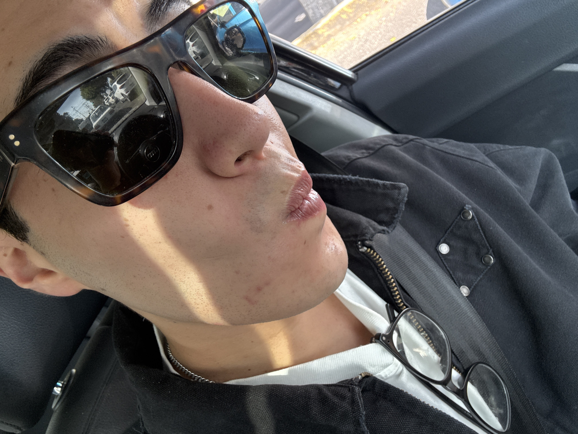 A close-up, angled shot captures a person inside a vehicle, seen from the mid-chest up. The individual, whose eyes are obscured by large, rectangular tortoiseshell-patterned sunglasses, is angled towards a window with their lips slightly pursed. Strong sunlight illuminates the scene, casting distinct shadows across their face, which shows light stubble, and clothing.
They are wearing a dark-colored jacket, possibly black denim or canvas, featuring a visible zipper and metal snaps, layered over a white t-shirt. A thin silver chain is visible around their neck. Tucked into the front of the jacket is a second pair of black-framed clear glasses.
The immediate surroundings suggest the interior of a car, with a dark car seat and a dark gray door panel visible. The reflections in the sunglasses provide a view of the outside environment, revealing several white vehicles (possibly vans or SUVs), trees, power lines, and what appears to be a street or road scene. A circular logo with indiscernible text is also reflected in the right lens of the sunglasses. The bright light and sharp shadows indicate a clear, sunny daytime. Given the context, this street scene is in Puebla, Mexico.