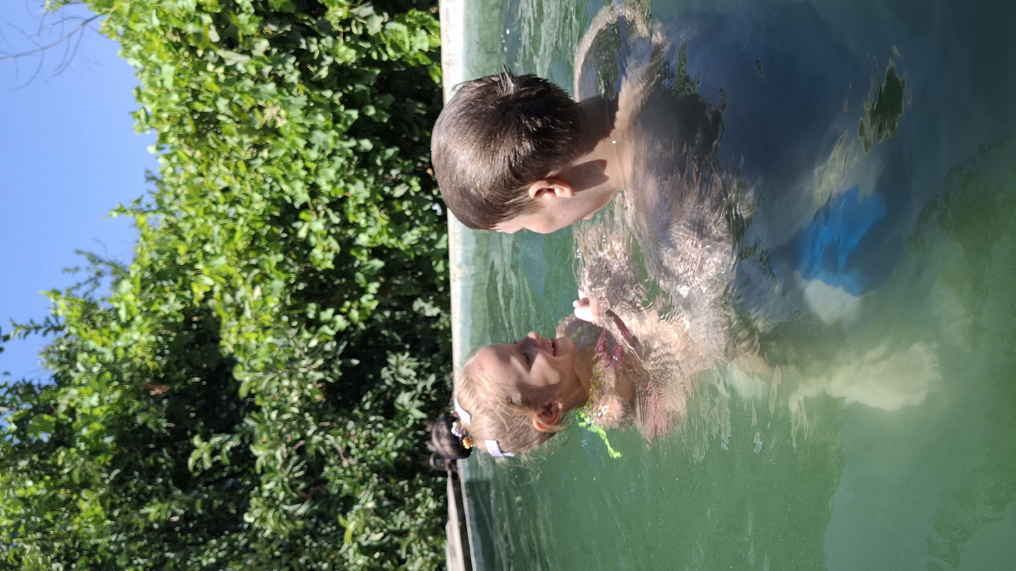 The image depicts two children in a swimming pool on a sunny day. The child on the left, a young girl with blonde hair adorned with white and pink bows, is smiling and looking at the child on the right. She is wearing a pink swimsuit. The child on the right, a boy with brown hair, is facing the girl. The water is green, and the pool appears to be an outdoor one surrounded by lush green foliage. The sky above is clear and blue. The location is stated to be in the city of Шұбарсу, Kazakhstan.