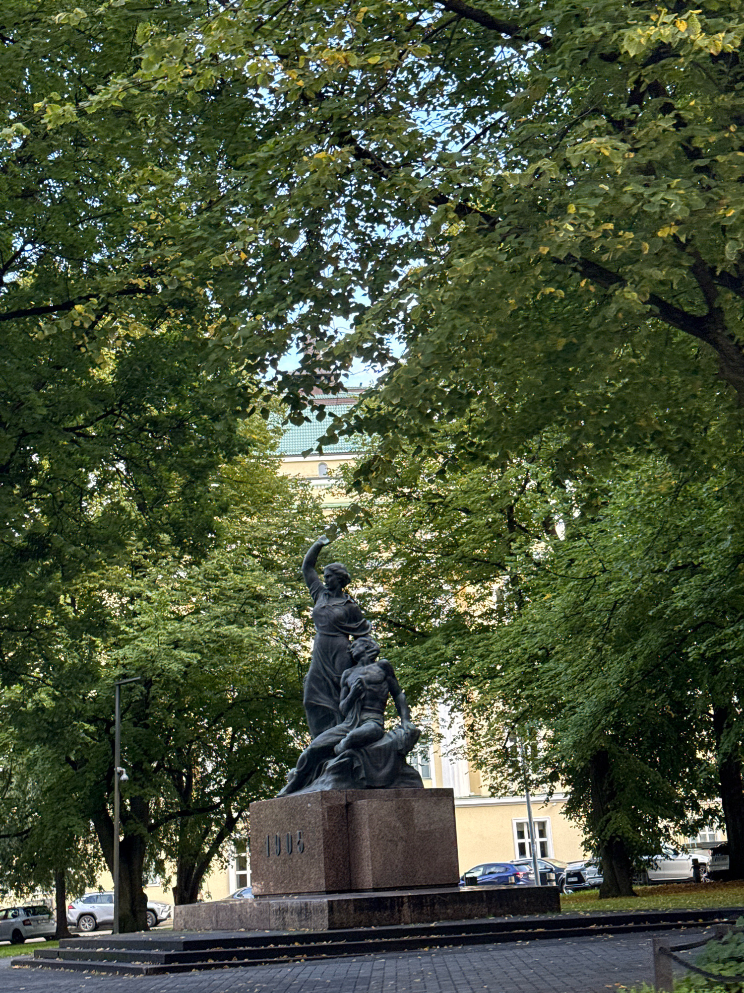 The image depicts the Hellemann Gate statue, also known as the "Victory of Light over Darkness," located in Tallinn, Estonia. The statue, made of bronze, stands on a granite pedestal inscribed with the year "1905." It features a female figure in a dynamic pose, reaching upwards with one arm, while a male figure is shown in a defeated or supportive posture at her feet. The statue is situated within a park-like setting, with lush green trees partially obscuring the background. In the distance, a building with a yellow facade and a green tiled roof is visible. Several cars are parked along the street in front of the building, and a lamppost with a security camera is located to the left of the statue. The ground in front of the statue is paved with dark tiles, and scattered fallen leaves suggest it might be autumn.