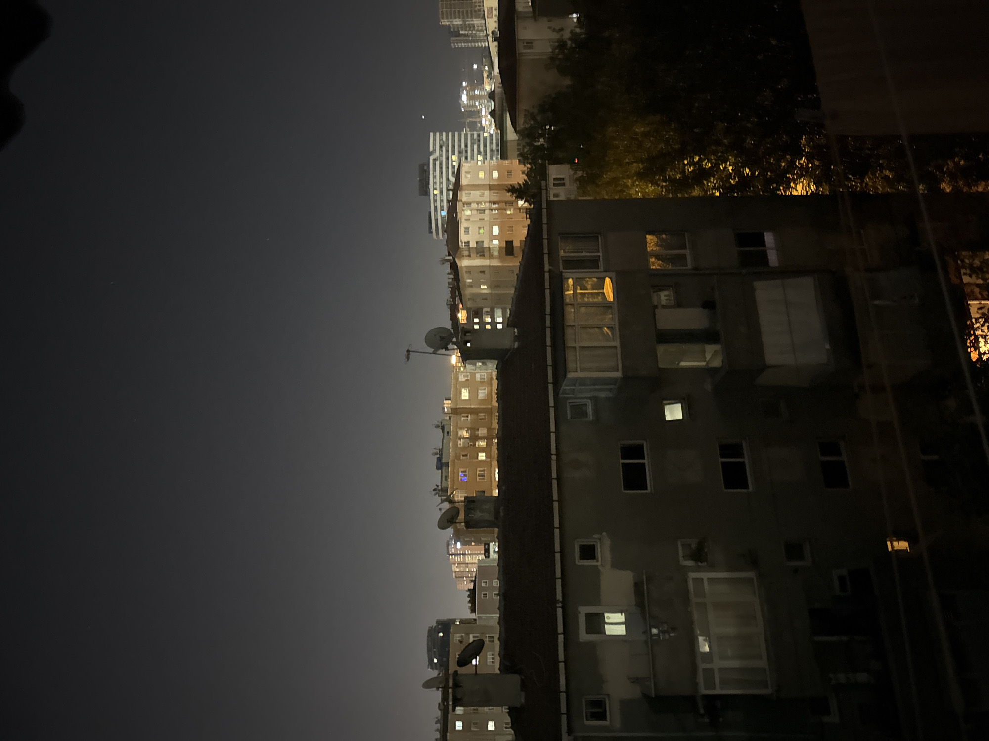 The image depicts a nighttime cityscape, likely from a residential building, overlooking other apartment blocks and a distant skyline in Gungoren, Turkey. In the foreground, a concrete building with several windows, some illuminated with warm light, is visible. The roof of this building is dark and sloped. Beyond this structure, several other apartment buildings of varying heights and architectural styles are scattered across the frame. Many of these buildings have windows lit up, suggesting occupancy. Some of these buildings feature satellite dishes on their roofs, common in urban areas. The background reveals a more distant urban landscape with taller buildings, including one with a distinct modern facade and numerous illuminated windows. The sky is dark and appears clear, with a faint haze near the horizon where city lights are most concentrated. The overall scene conveys a sense of urban life at night, with the glowing windows of residential buildings creating a pattern against the dark sky. There are no people or specific actions visible in the image. The environment is predominantly built-up urban infrastructure.