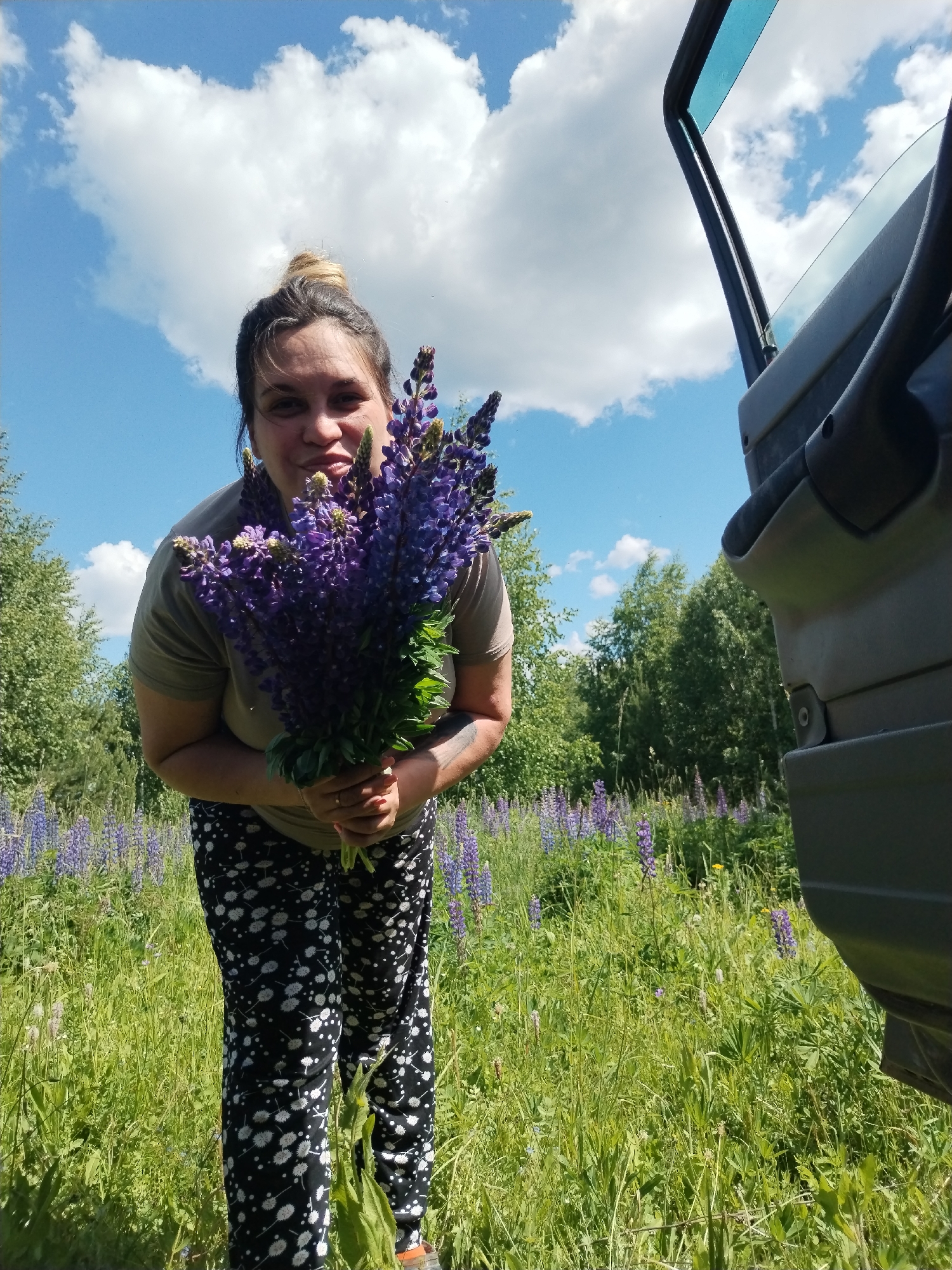 A woman stands in a field of purple lupine flowers, holding a large bouquet of the same flowers. She is wearing a plain green t-shirt and black leggings with a white floral pattern. The sky above is blue with fluffy white clouds. To the right of the woman is the open door of a dark-colored vehicle. The scene appears to be in a rural area, possibly Zipunovo, Russia, given the presence of the lupine field and the natural environment. The woman is looking towards the camera with a slight smile. The overall impression is a pleasant outdoor scene on a sunny day.