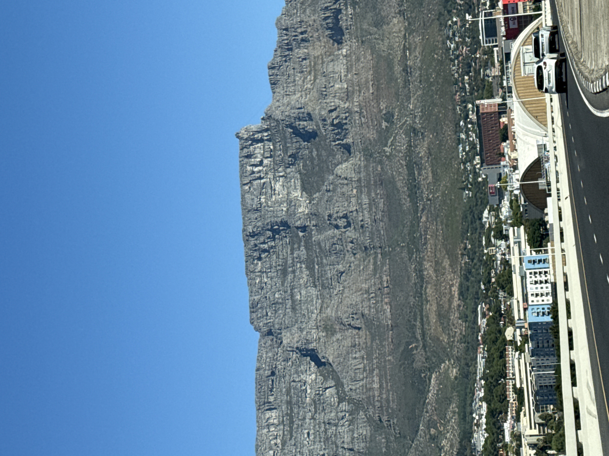 The image depicts Table Mountain, a prominent landmark overlooking Cape Town, South Africa. The mountain's steep, gray rock face dominates the scene, contrasting sharply with the clear blue sky. Below the mountain lies a dense urban landscape consisting of buildings, roads, and vegetation. A highway with moving vehicles is visible alongside the urban area, indicating proximity to the city center. The overall impression is a combination of natural grandeur and urban development.