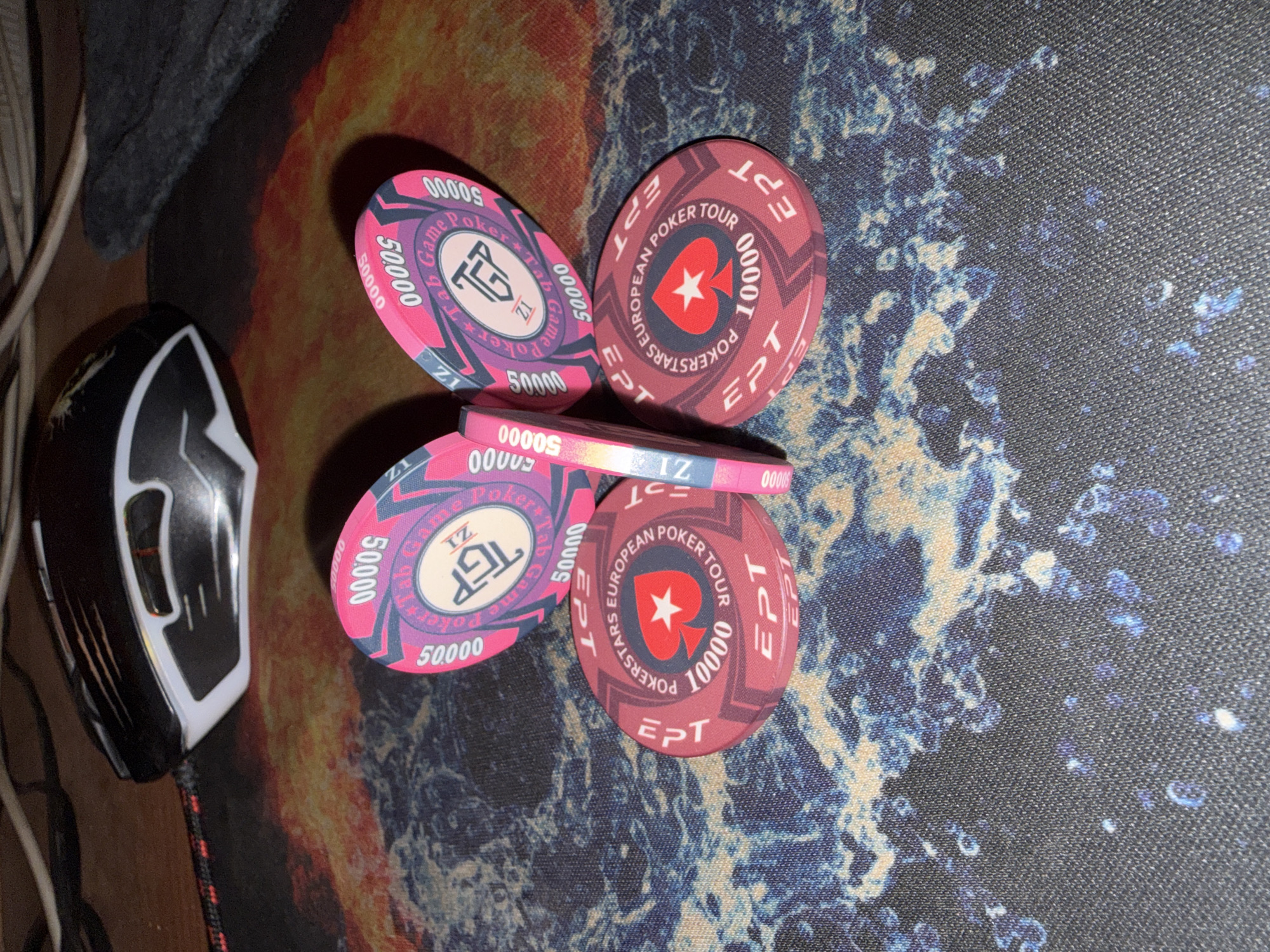 The image shows a black and white computer mouse next to a small stack of pink and red poker chips. The poker chips appear to be arranged in a circular fashion. The poker chips are resting on a multicolored mousepad. The scene appears to be located indoors, on a desk or similar surface. The text printed on the poker chips include terms such as “European Poker Tour”, "Pokerstars", "Game Poker Tab”, “EPT” and numerical values such as “10000”, “50000”. There is no evidence to indicate that the image was taken in Novyi Menzelyabash, Russia.