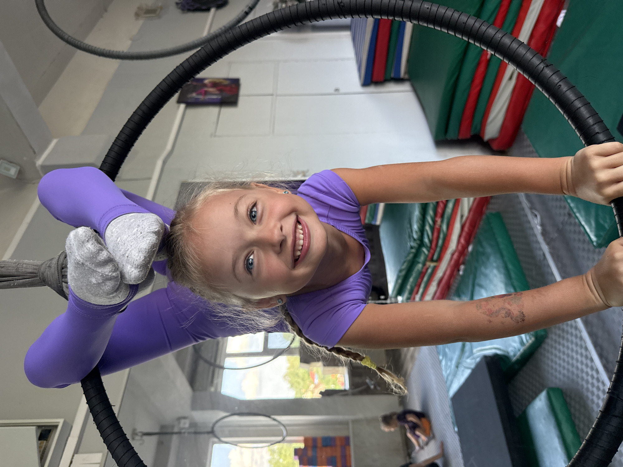 A young blonde girl with blue eyes, wearing a purple top and purple leggings with grey socks, is hanging upside down from an aerial hoop. Her hair, braided with a yellow tie, is also hanging downwards. She is smiling brightly at the camera, with a happy and confident expression. Her left leg is bent, with her foot resting against the hoop, while her right leg is extended straight. Her arms are extended, gripping the black hoop.
The background suggests an indoor gymnasium or acrobatic studio. Stacked mats of various colors, including green, red, and white, are visible on the right side. The walls are painted a light color, and there are windows that allow natural light into the space. A few other people are faintly visible in the background, one of them possibly a child.
The overall impression is one of a child enjoying an acrobatic activity. The lighting appears to be natural daylight, suggesting the activity is taking place during the day. The setting is an athletic facility, likely for gymnastics or aerial arts. There is a visible tattoo on the girl's right forearm. No discernible text is visible in the image. The context provided indicates the location is Dnipro, Ukraine.