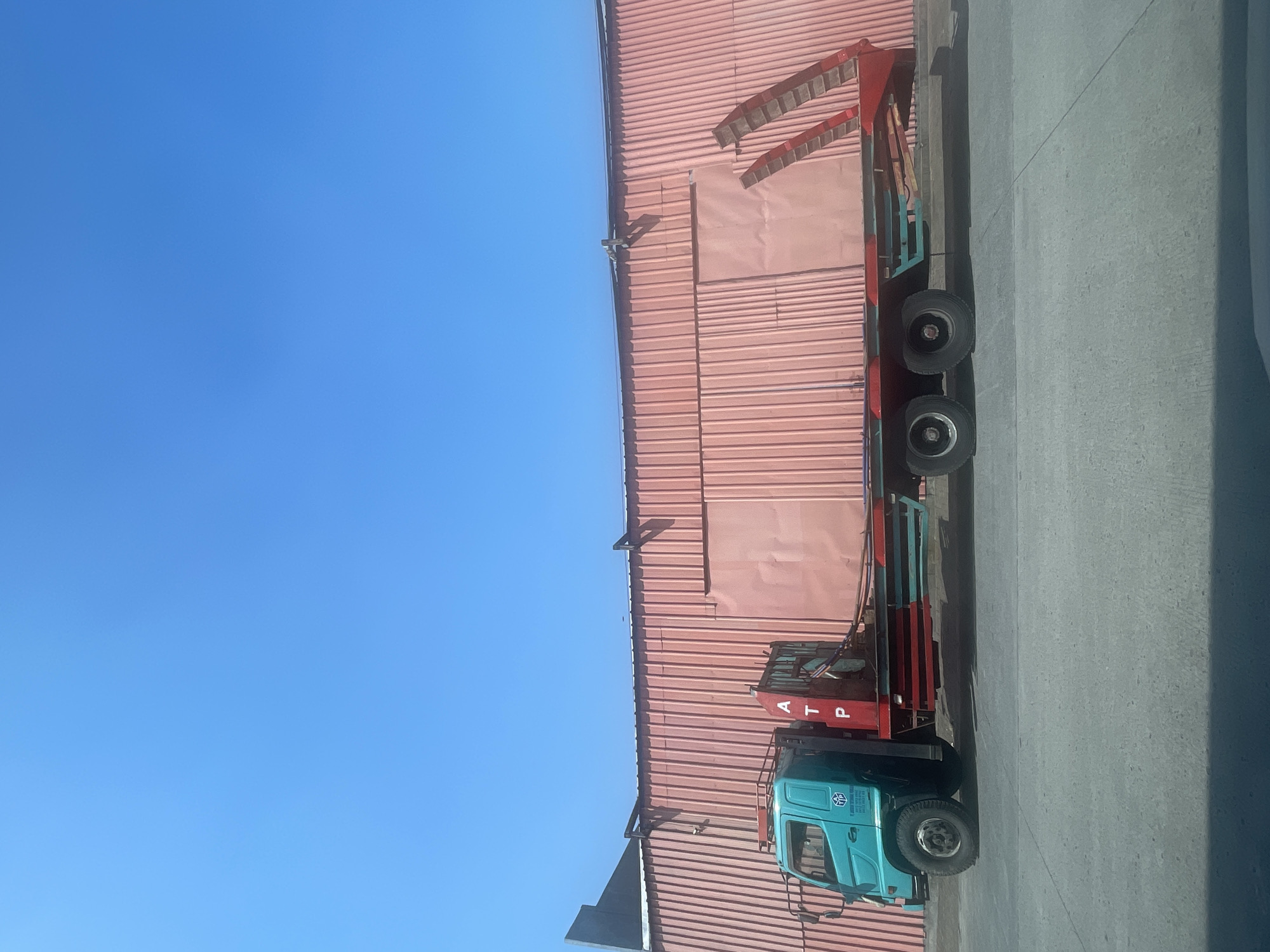 A teal and red flatbed truck is parked on a concrete surface in front of a large, pink corrugated metal building. The truck has a teal cabin with a blue logo and "ATP" written vertically in red letters on a support structure behind the cabin. The flatbed is empty and has red and teal accents along its sides. The building has a series of vertical panels and appears to be a warehouse or industrial structure. The sky is clear and blue. The scene is outdoors, likely in a commercial or industrial area. The lighting suggests it is daytime, with the sun casting shadows.
