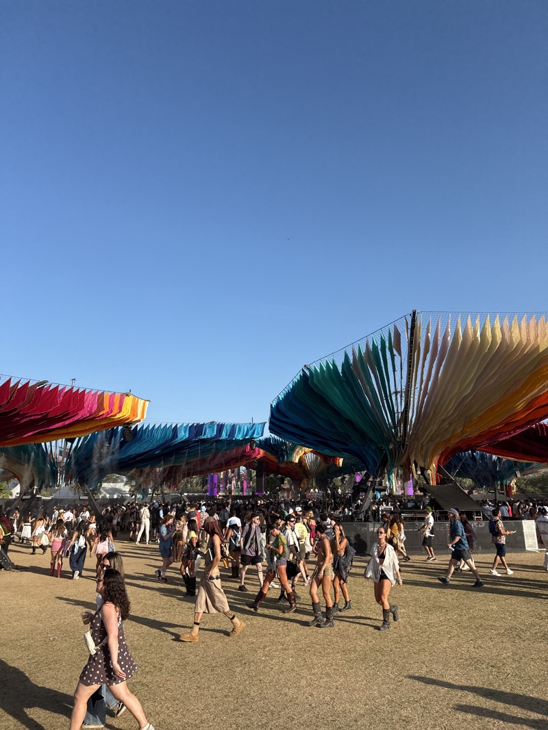 This image captures a vibrant, sunny outdoor festival scene in Indio, United States. The setting is a large, open field with dry, sparse grass, under a clear, bright blue sky.
The primary subjects are a large crowd of people, predominantly young adults, walking across the field. They are dressed in diverse, casual attire, often typical of festival fashion, including shorts, dresses, light tops, and boots. Many individuals are engaged in walking and moving through the event grounds, with some carrying bags or drinks.
Above the crowd, large and elaborate art installations or decorative canopies stretch across the sky. These structures are made from numerous vertical strips of fabric, arranged in sweeping, fan-like or wave-like patterns, displaying a wide spectrum of colors, including reds, oranges, yellows, greens, and blues, creating a rainbow effect. In the background, a stage or elevated platform is partially visible amidst the crowd and structures.
The activity taking place is a large-scale outdoor gathering, likely a music or arts festival, where people are moving between different areas.
Notable details include the time of day, which is bright daylight, indicated by the clear blue sky and strong shadows cast by the people and structures on the ground, suggesting mid-morning to late afternoon. The weather is sunny and clear, with no clouds visible. The colorful fabric installations serve as prominent visual cues, characteristic of many desert festivals. The overall mood appears lively and relaxed. There is no visible text in the image.