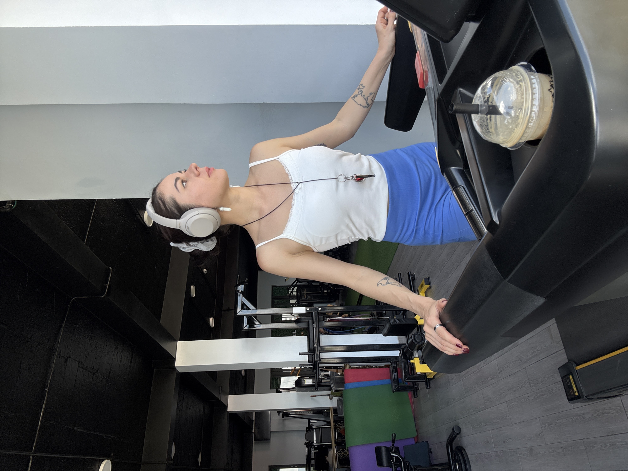 The image shows a young woman on a treadmill in a gym. She is wearing a white tank top, blue shorts, and headphones. She is looking to the side with a determined expression. In the background, there are various gym equipment, including weight machines and mats. The gym appears to be well-lit and spacious. A cup with a beverage is placed in the treadmill's cup holder. The overall scene suggests a focused and active individual engaged in a workout. The location is not specified as Da Lat, Vietnam.
