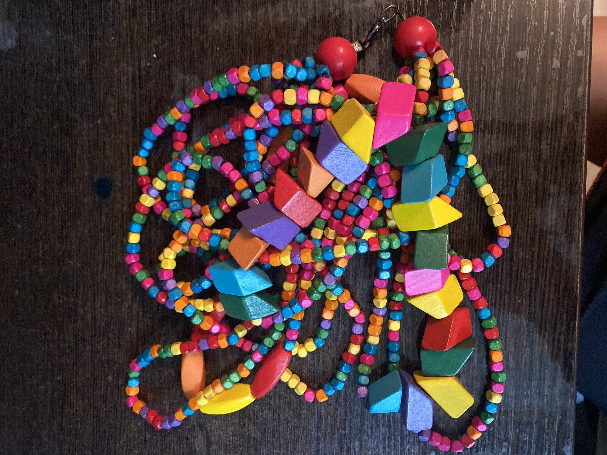 The image shows a close-up, overhead view of a colorful, wooden beaded necklace, likely handmade, lying on a dark wooden surface. The necklace is long and features a variety of bead shapes and sizes, including small, cylindrical beads in multiple bright colors like red, orange, yellow, green, blue, and purple. Interspersed among the smaller beads are larger, geometric wooden pieces in vibrant hues of yellow, red, orange, purple, blue, and green. These larger pieces are also varied in shape, including diamond-like facets and elongated oval shapes. The necklace is coiled and draped loosely, creating a complex pattern. Towards the bottom right of the image, the necklace appears to have a closure mechanism with two red spherical beads and a metal clasp. There is no indication of the necklace's location in Ivanteyevka, Russia, or any other specific city. The focus is solely on the object itself.