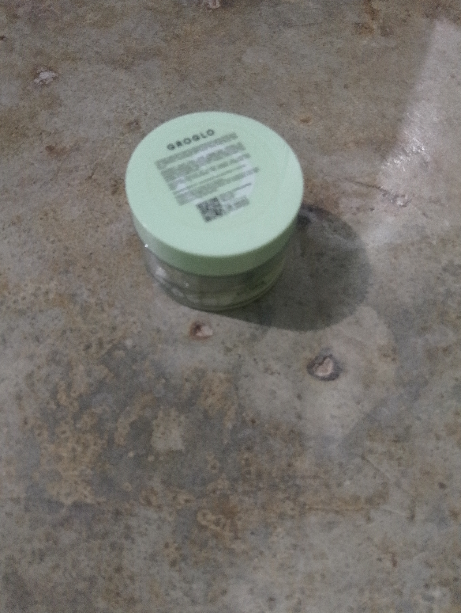 A light green, circular container with white text and a QR code is placed on a rough, textured, concrete-like surface. The text on the container appears to be in English, with "GROGLO" clearly visible at the top. The container is translucent, with some of its contents visible inside. The surface it rests on has variations in color and texture, with small flecks and darker patches. The lighting suggests an indoor or shaded outdoor environment, casting a soft shadow to the right of the container. No people are present in the image, and no specific location in Serbelawan, Indonesia, can be identified from the visual information provided.