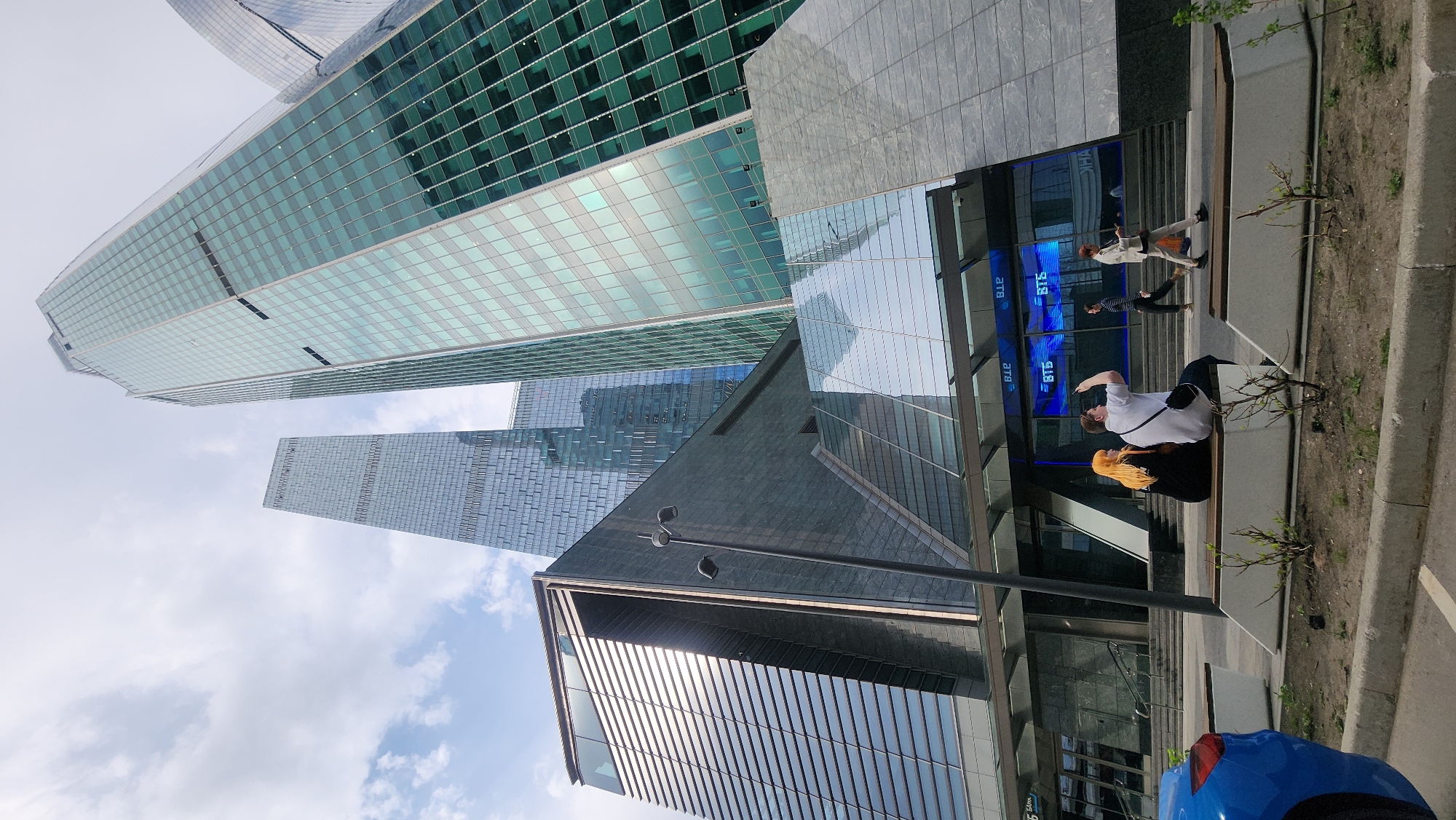 A low-angle, daytime shot captures several modern skyscrapers in Moscow, Russia, combined with ground-level elements like people, street furniture, and part of a vehicle.
In the background, two prominent skyscrapers feature glass facades. One has a green tint and a grid-like pattern, while the other has a gray tint and a similar grid. The sky appears partly cloudy.
Closer to the viewer are architectural details of another building, including reflective glass and concrete surfaces. A slender streetlight is visible.
On the ground, a couple is seated on a concrete bench. One person is wearing a white shirt and has a dark bag, seemingly taking a photo with a phone. The other person is wearing a dark outfit with blonde hair. Beyond them, two individuals are walking.
Part of a blue car is visible in the lower right corner, along with a paved sidewalk and sparse vegetation. The image conveys a sense of urban life juxtaposed with modern architecture in Moscow.