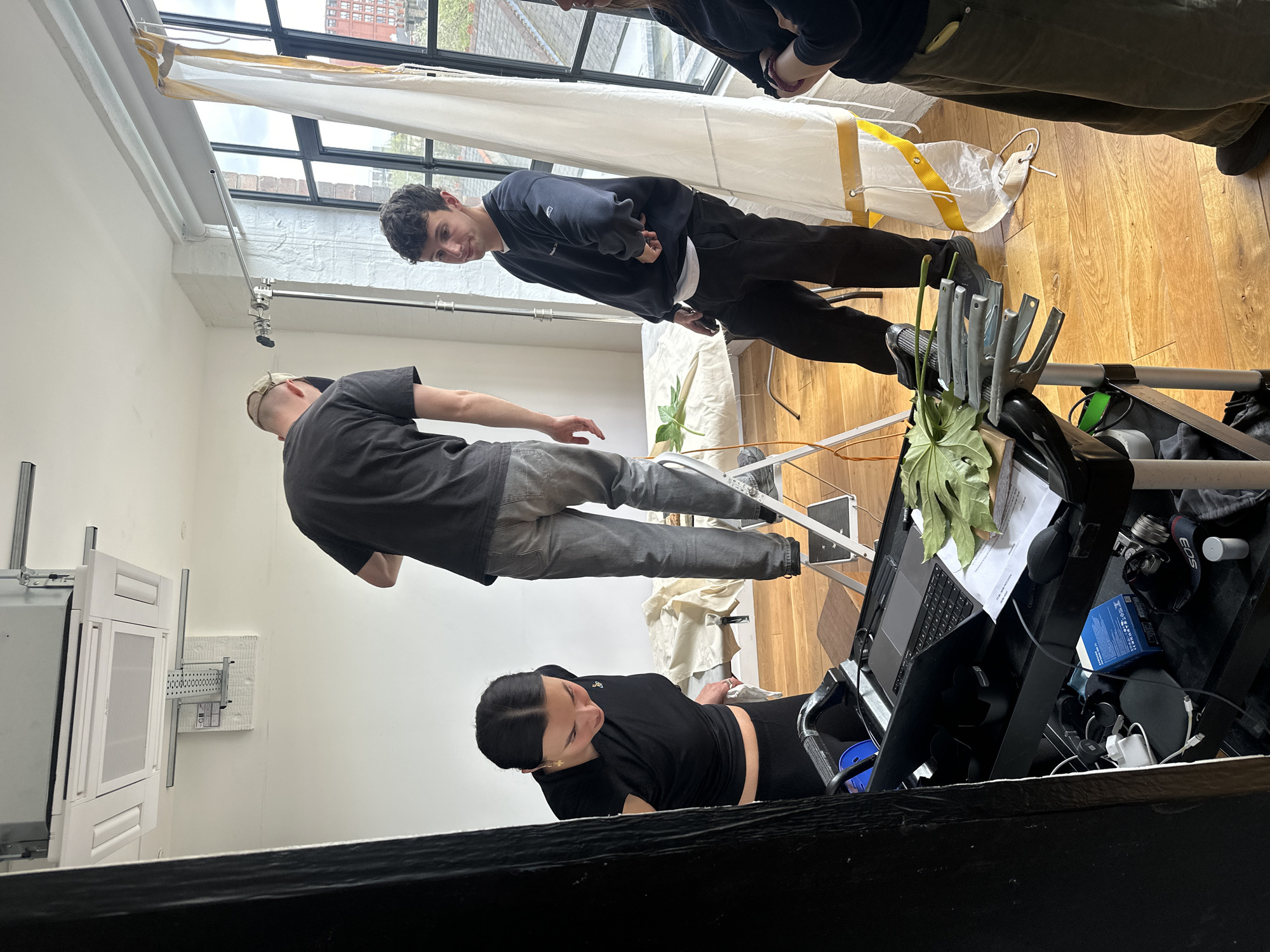 The image depicts an indoor scene, likely a photography or film studio, where three individuals are engaged in a production session. On the right, a young man with dark, curly hair, wearing a black sweatshirt with "MBUK" visible on it and dark trousers, stands observing, looking towards the left with a slight smile. Towards the center, another man, wearing a black t-shirt, grey jeans, and a baseball cap, has his back to the camera and is leaning forward, possibly adjusting equipment or a prop. On the far left, a woman with dark hair pulled back, dressed in a black t-shirt and dark leggings, is seated or leaning over a black rolling cart, intently looking down at a MacBook laptop screen, suggesting she is monitoring the shoot or working on visuals.
The setting is brightly lit, primarily by natural light streaming through large windows with black frames on the right, which reveal an urban backdrop of brick buildings and an overcast sky, characteristic of London. A large white fabric light modifier with yellow straps is positioned near the window. The floor is made of light-colored wooden planks, and the walls are white.
Numerous pieces of studio equipment are visible. On the black cart near the woman, a MacBook laptop, a leafy plant, camera accessories including an "EOS" lens cap, a blue box with foreign text, and various cables are arranged. In the foreground, metal clamps are attached to a stand, and another leafy plant is visible. A c-stand with a boom arm extends from the wall. On the far left wall, a white cabinet-like structure with a metal grate and support rails is mounted. The scene conveys a focused and professional creative atmosphere during daytime.