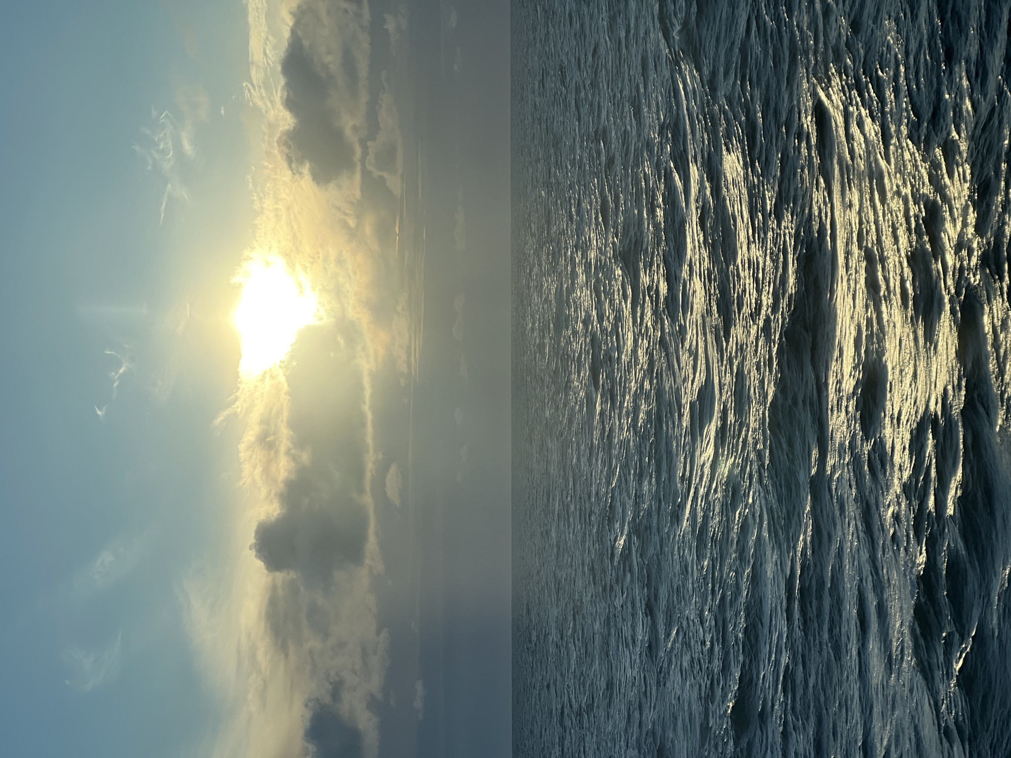 The image displays a vast expanse of choppy, dark blue ocean under a sky transitioning from clear light blue at the top to overcast with bright sunlight peeking through the clouds near the horizon. The sun is a brilliant, blown-out white, partially obscured by a band of clouds, casting a warm, golden light that reflects off the surface of the water. The waves are clearly defined, with highlights and shadows creating a textured appearance. The overall scene evokes a sense of natural beauty and vastness. There are no people, structures, or specific landmarks visible that would indicate a location within the city of Unknown, Poland, or any other specific geographical area. The environment is solely the open sea and sky.