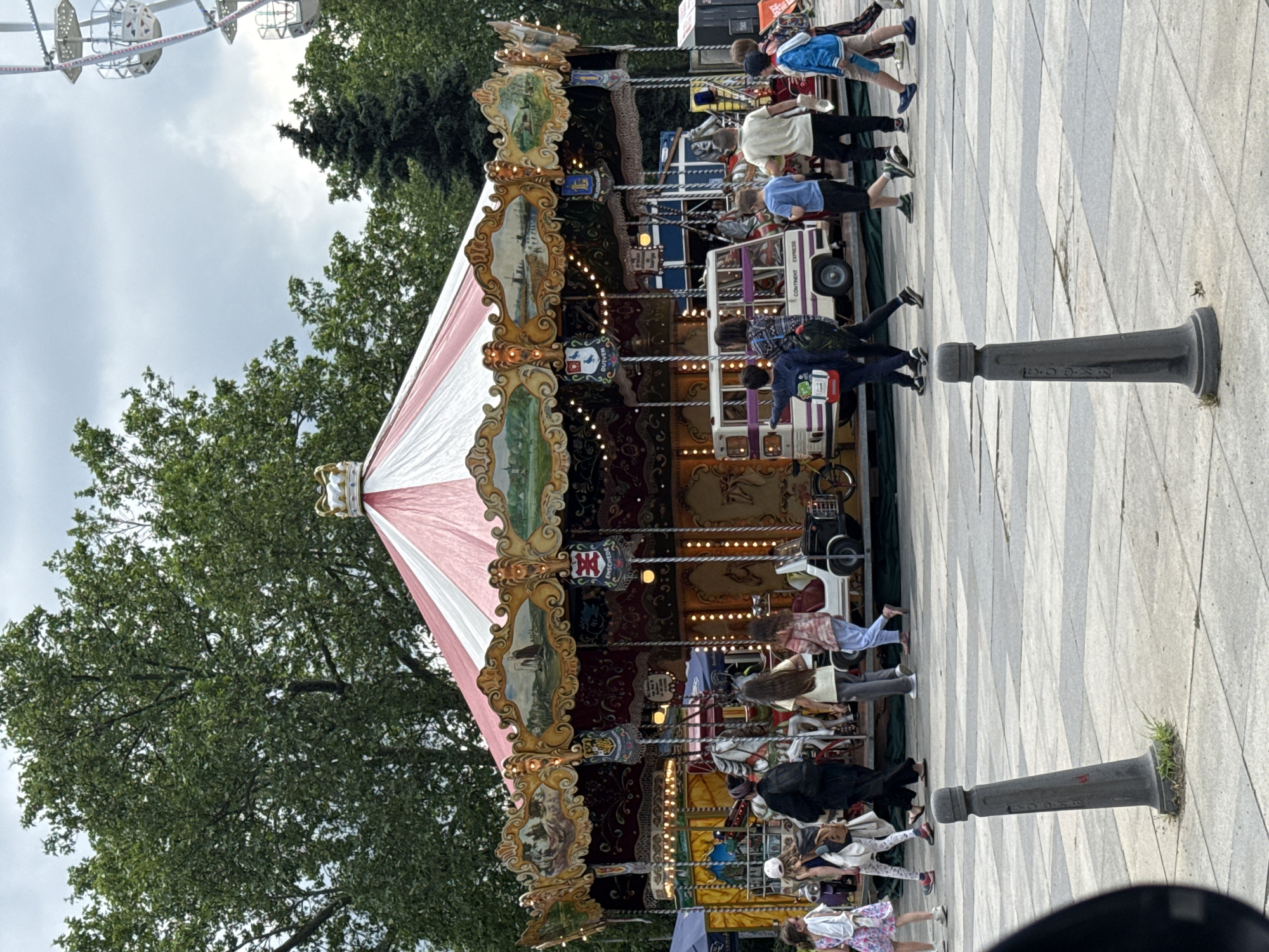 This image captures a vibrant scene at an outdoor amusement area, likely a park or fairground in Sofia, Bulgaria, on an overcast day. The central focus is a highly ornate, traditional carousel with a distinctive white and pink striped canopy crowned by a golden ornament.
The carousel is bustling with activity, featuring multiple children and adults. Children can be seen riding various carousel animals and vehicles, including a small bus-like carriage clearly labeled "CONTINENT EXPRESS." Adults are standing around the perimeter, some observing the riders, others possibly waiting their turn or accompanying children. The carousel itself is richly decorated with golden accents, intricate painted panels depicting landscapes, and heraldic crests, some of which feature a double-barred cross symbol, though the text on them is largely indecipherable. Small, warm lights adorn the carousel's structure, suggesting an inviting atmosphere even under cloudy skies.
In the background, lush green trees provide a natural setting, and the upper left corner reveals part of a large Ferris wheel, further confirming the location as an amusement venue. The ground is paved with a distinct gray and white geometric pattern. Two black bollards are visible on the right side of the frame; one of them has partially visible text, appearing to be "P.D.O.S." or similar. The overall ambiance is relaxed and lively, indicative of recreational enjoyment. The cloudy sky suggests daytime, possibly late morning or afternoon, with diffused lighting.
