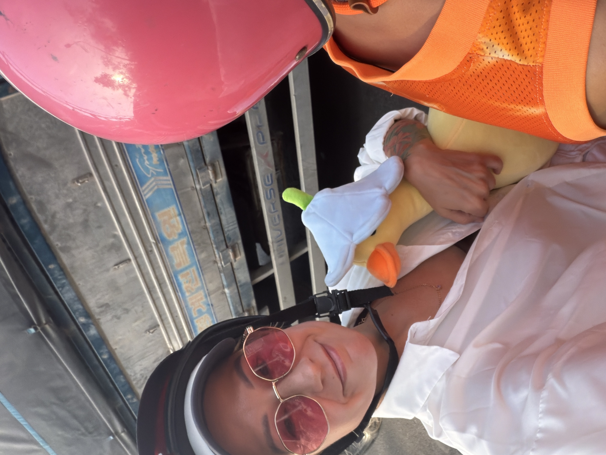A woman in a white shirt and a black helmet with a pink tint to her round sunglasses smiles while holding a plush toy duck. Her left arm, adorned with a tattoo, rests on the duck. To the right of the frame, a person wearing a bright orange mesh garment and a pink helmet is partially visible. They appear to be on a motorbike, with the side of a vehicle with Korean writing visible in the background. The scene suggests a warm, outdoor environment, possibly on a street in South Quy Nhon Ward, Vietnam.