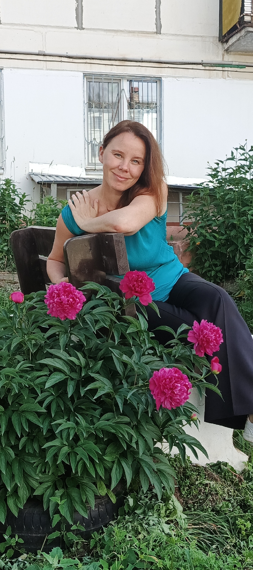 An outdoor medium shot captures a woman sitting on a dark wooden bench in front of a building with white walls and barred windows. She has medium-length brown hair, is wearing a bright teal tank top and dark pants, and is leaning on the back of the bench. In the foreground, vibrant pink peonies are in bloom. The scene appears to be in a residential area, possibly in Chaikovsky, Russia, given the architectural style of the building and the presence of peonies, which are common in Russian gardens.