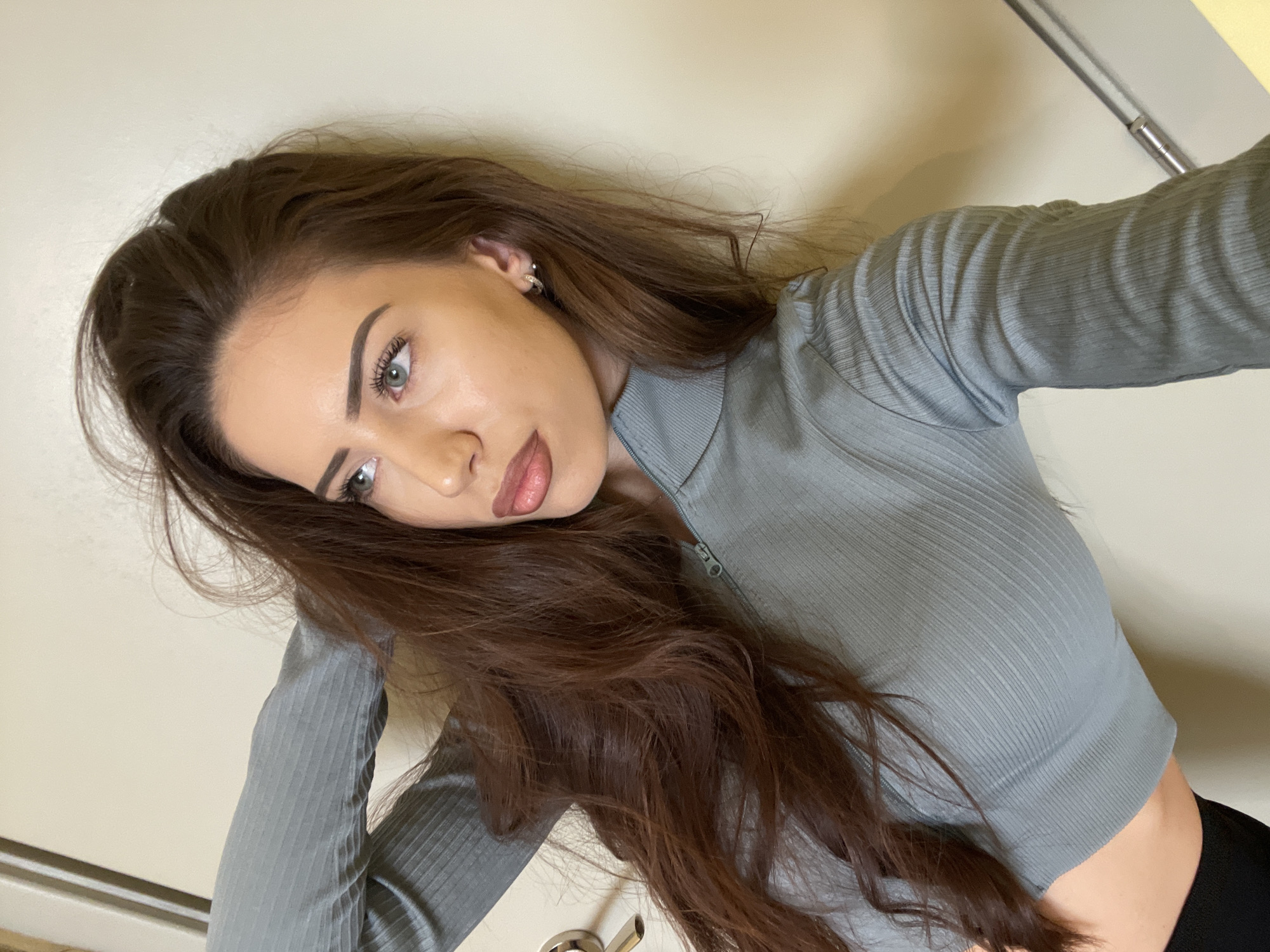 A young woman with long, brown hair is posing for a selfie indoors. She is wearing a light blue, long-sleeved, ribbed shirt with a zipper at the neckline. She has fair skin, defined eyebrows, and appears to be wearing makeup. Her eyes are light-colored. She is wearing a small earring in her left ear. The background is a plain, off-white wall with a door frame visible in the upper right corner, with silver hardware. There is no indication of the location being Veliko Tarnovo, Bulgaria in the scene.