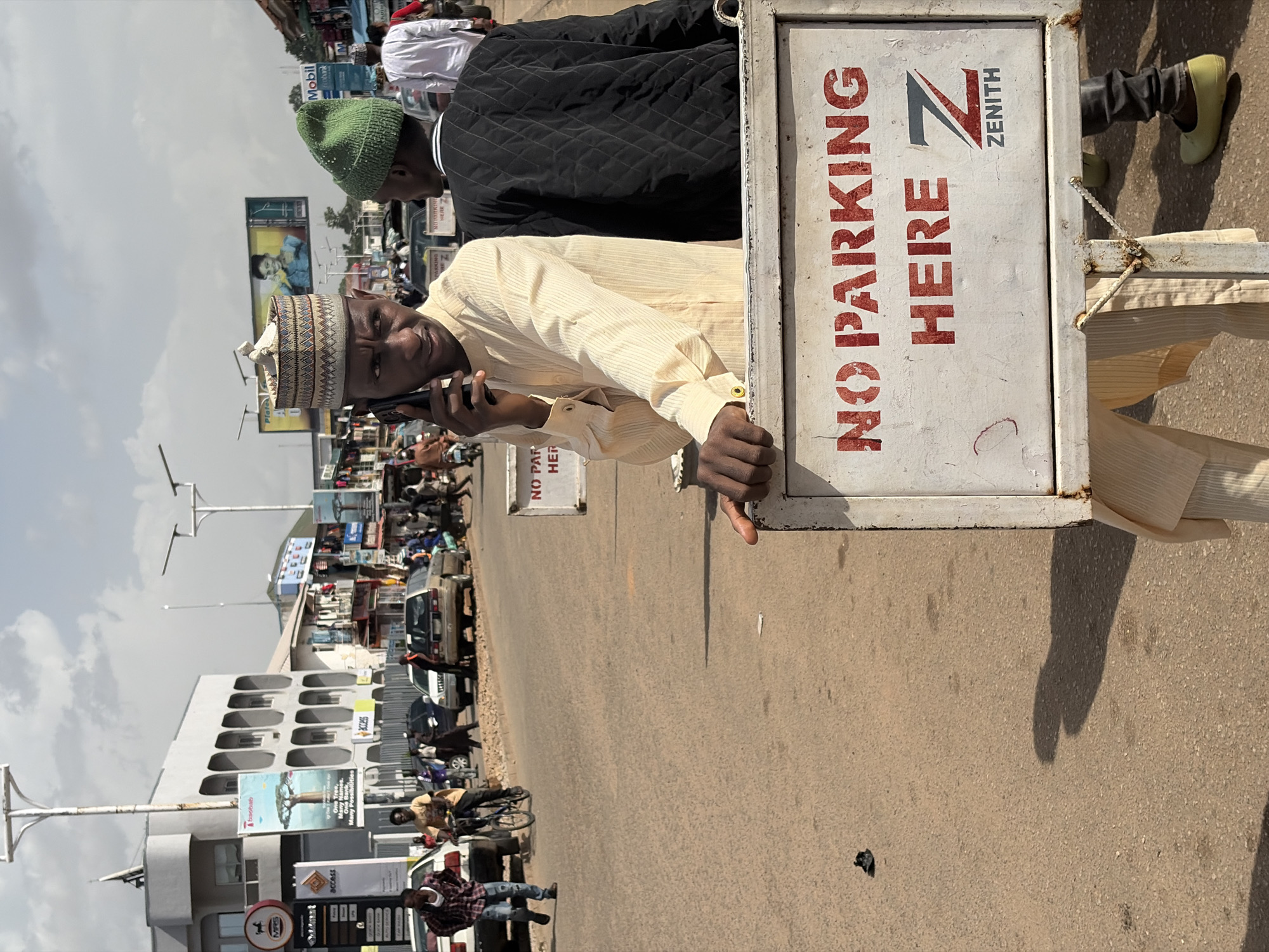 This is a vertical, eye-level shot captured in Jos, Nigeria. The scene appears to be a bustling street or marketplace, with a mix of people, vehicles, and buildings. The sky is overcast, suggesting a cloudy day.
The foreground is dominated by a man on the right side of the frame, wearing a light-colored shirt and a traditional cap. He is holding a mobile phone to his ear, engrossed in a conversation. Partially obscuring him is a sign that reads "NO PARKING HERE" in bold red letters, with the "ZENITH" logo visible. To the left of this man, another man wearing a black quilted jacket and a green beanie is visible.
The background reveals a busy street with various vehicles, including cars and motorcycles, parked and moving. There are also buildings with storefronts and advertisements, one of which shows "Mobil". Signs for "access" and "Cobank" are also visible, along with another "NO PARKING" sign. The overall impression is one of a lively, everyday scene in an urban Nigerian environment. The people seem engaged in their daily activities, with no overt signs of distress or unusual events. The lighting suggests it is daytime.