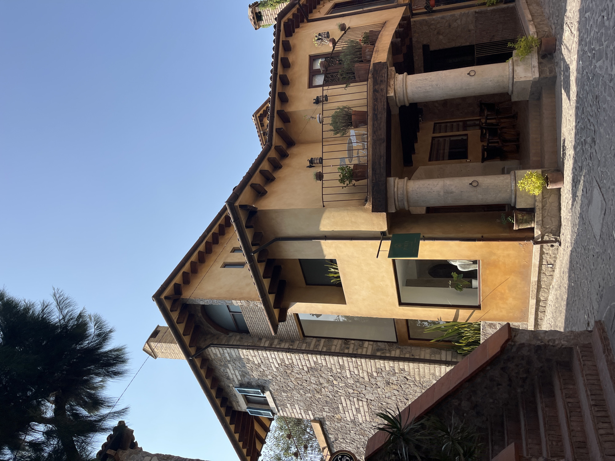 The image captures a sunlit architectural scene in Domingo Arenas, Mexico, featuring multi-story buildings with a blend of stone and stucco facades. The architectural style exhibits elements of Mediterranean or Tuscan design, characterized by exposed beams, tile roofs, and arched balconies with potted plants. Stone steps and pathways delineate the foreground, while the clear blue sky offers a backdrop to the buildings. The scene appears tranquil, devoid of human activity, and emphasizes the architectural details and natural light.