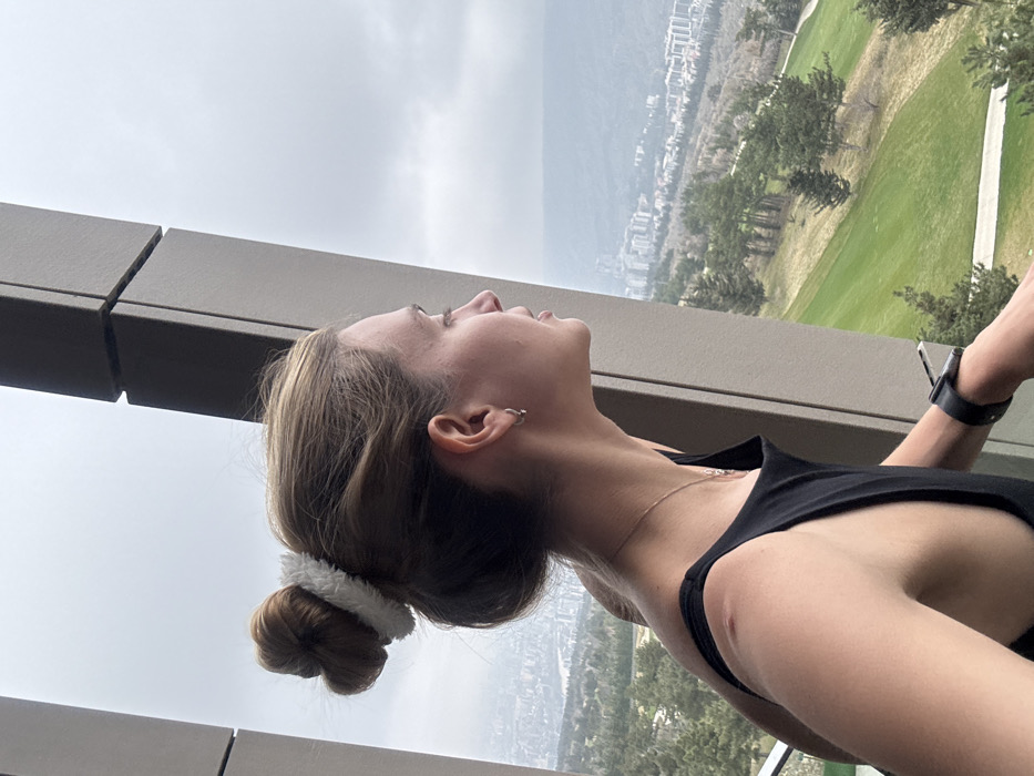 A female individual is depicted in profile, facing towards the right side of the frame, positioned on what appears to be a balcony or observation deck of a modern building. Her light brown hair is pulled into a bun, secured by a white, fluffy scrunchie. She wears a black sleeveless top, a thin gold-colored necklace with a small pendant, and small hoop earrings in her visible left ear. A dark-colored smartwatch is present on her left wrist. A small reddish mark is visible on her right shoulder. Her right hand rests on a beige-grey angled architectural beam that forms part of the structure. The background features an expansive panoramic view of the Tbilisi Hills Golf & Residences area in Tbilisi, Georgia. In the mid-ground, a green golf course with manicured fairways and scattered pine trees is visible. Beyond, the cityscape of Tbilisi extends across undulating hills, partially obscured by an overcast sky.