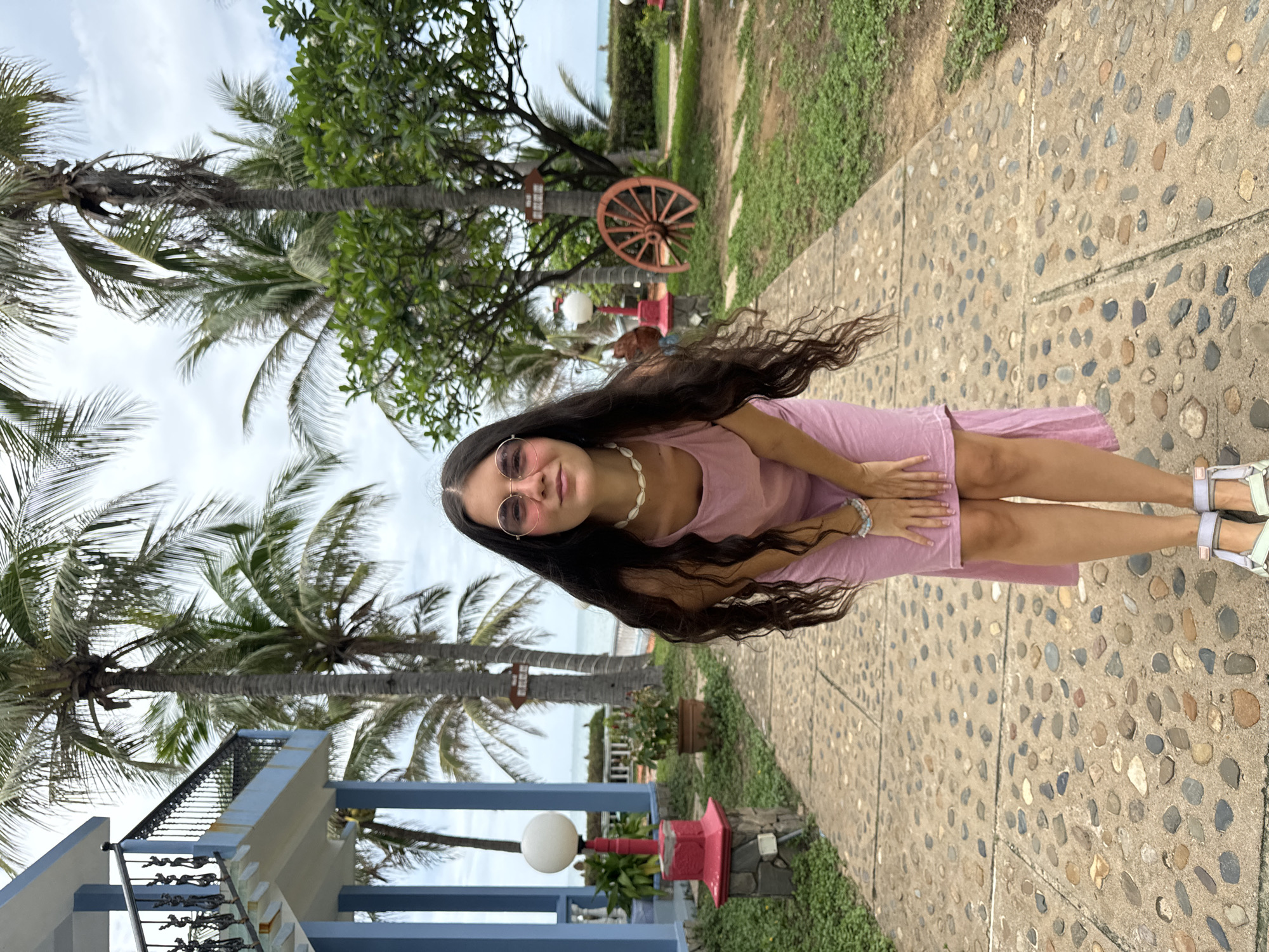 A young woman is featured in an outdoor, tropical setting, likely a resort or hotel garden in Mũi Né, Vietnam. She stands on a paved path made of concrete embedded with various colored pebbles, looking towards the camera with a slight smile. She has long, dark, wavy hair that flows over her shoulders and down her back. She is dressed in a light pink sleeveless dress, a white shell necklace, and pink-tinted round sunglasses. Her hands are clasped gently in front of her, and she wears light-colored sandals.
The background is dominated by numerous tall palm trees with green fronds, suggesting a lush, warm environment. On the left side of the frame, a modern building with blue and white railings is partially visible. Further into the background, to the right of the woman, there's a rustic, reddish-brown wagon wheel leaning against a tree trunk, accompanied by an outdoor lamp with a spherical white shade and some potted plants. Beyond the trees, a glimpse of the sea or a large body of water can be seen under a cloudy, overcast sky. The ground alongside the paved path is covered with green grass and other foliage. The scene captures a relaxed moment during daytime. No legible text is visible.
