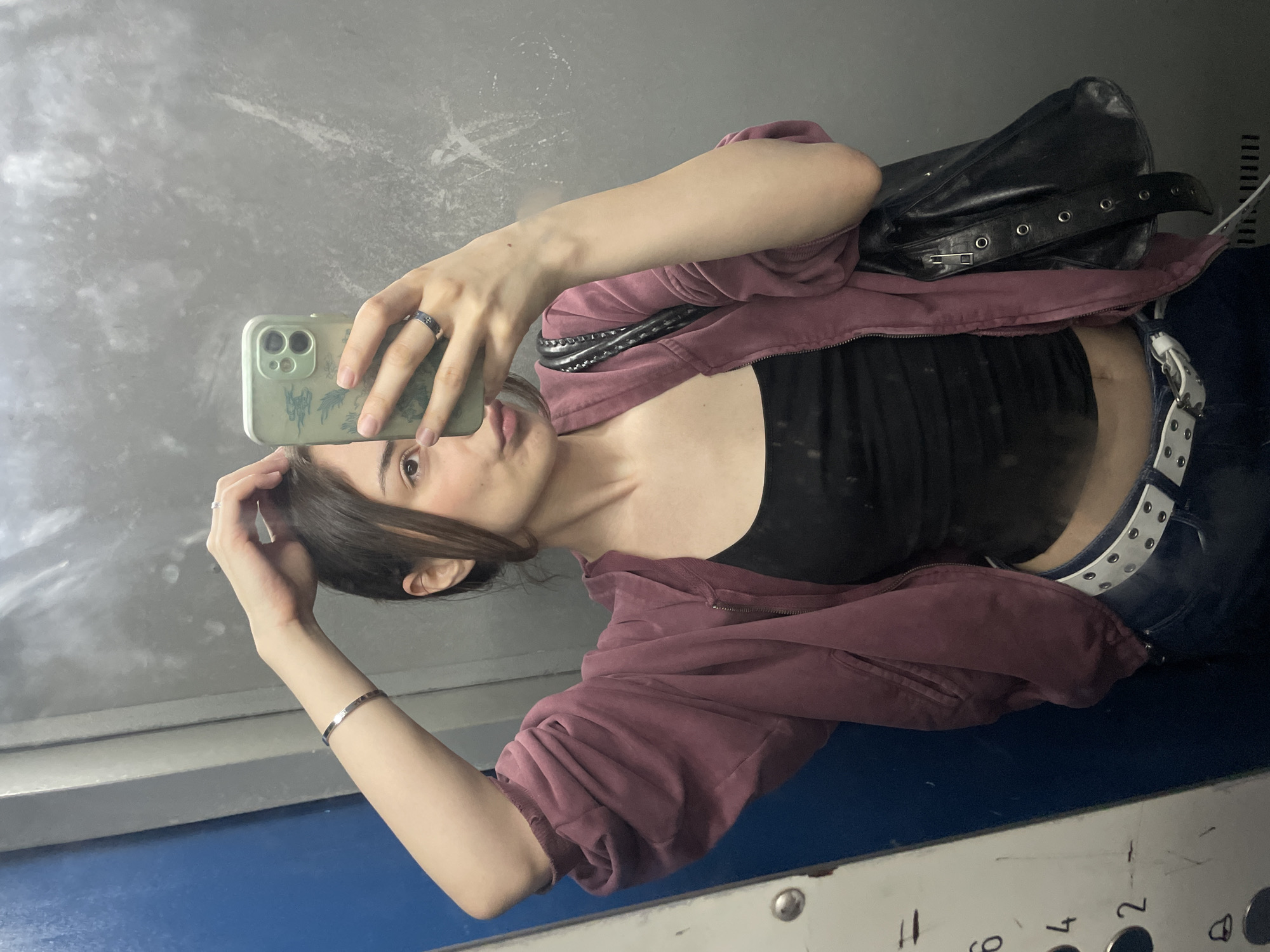 A young woman is taking a mirror selfie in an elevator. She is wearing a dark tank top, a dark purple zippered hoodie, and dark jeans with a white belt. She has a black purse slung over her right shoulder and is wearing a silver bracelet on her left wrist and a ring on her right index finger. The elevator walls are made of gray, textured material, and there is a blue door on the left side of the frame. A panel with numbers indicating elevator floors is visible on the left side. The background is slightly out of focus, suggesting the photo was taken in motion or with a shallow depth of field. The scene is indoors, in a confined space that appears to be an elevator.