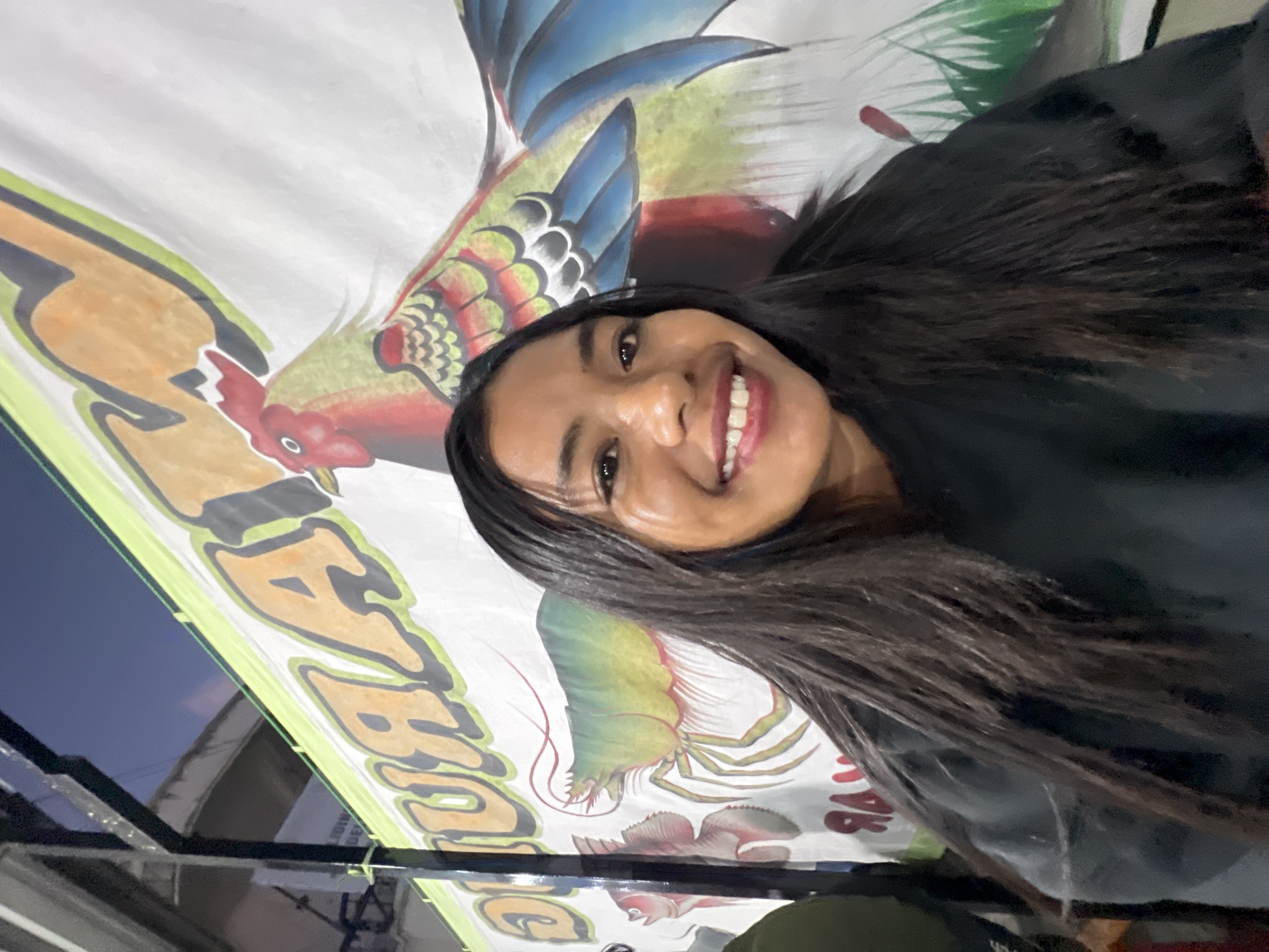 A young woman with long, dark hair smiles at the camera in a selfie taken in front of a brightly colored banner. The banner features illustrations of a rooster, a shrimp, and possibly a fish, with large, stylized lettering. The scene appears to be outdoors, with a glimpse of a dark sky and some structural elements suggesting a market or food stall. The woman is wearing a dark-colored top. The background banner has text that appears to read "WARUNG" which translates to "stall" or "eatery" in Indonesian. The artwork on the banner is consistent with typical decorations found at food stalls in Denpasar, Indonesia, particularly those specializing in seafood or poultry.