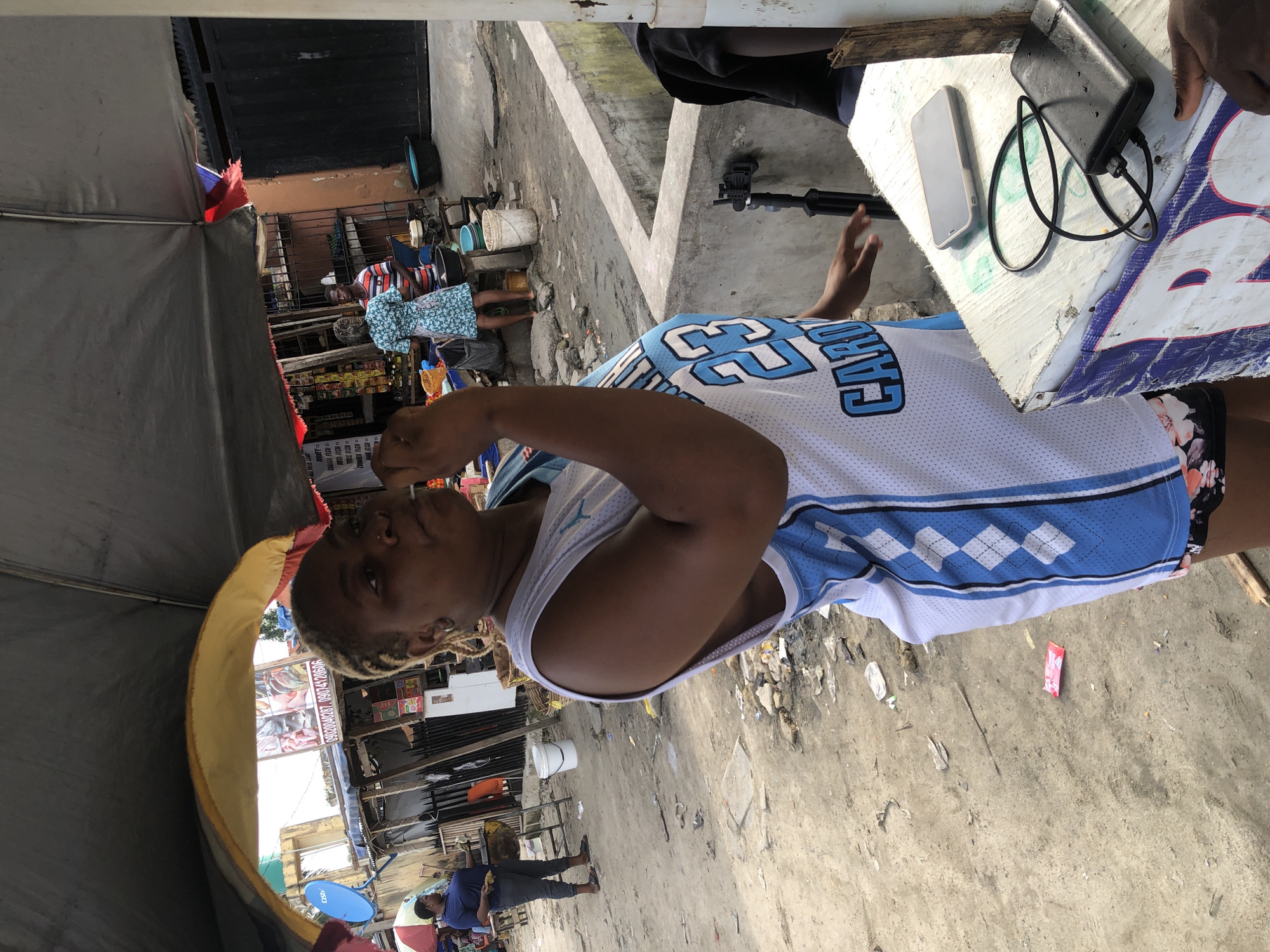 A medium shot, street level photograph captures a person of African descent wearing a white and blue basketball jersey with the number "23" on it, seemingly eating and looking directly at the camera. They are standing near a table with a cell phone, a charger, and a power bank on it. The background shows a busy outdoor market in what appears to be Ado, Nigeria. Other people can be seen in the background, along with market stalls and various objects scattered on the ground. The overall lighting suggests a sunny day.