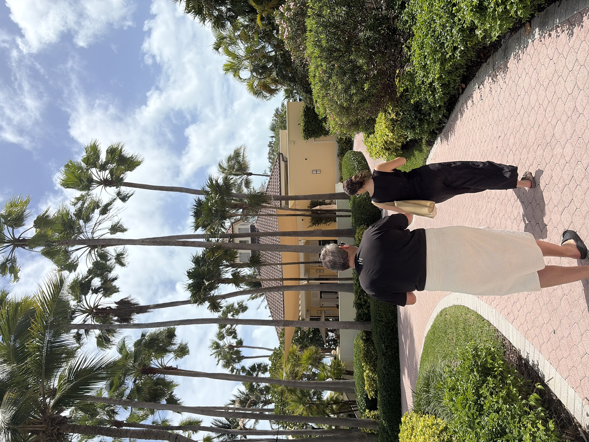 The image depicts an outdoor scene with two people walking along a brick path. A taller man in a black shirt and light-colored shorts walks ahead of a shorter woman wearing a black jumpsuit and carrying a beige bag. The path is bordered by lush green bushes and trees on one side and a row of tall palm trees on the other, leading to a tan-colored building in the background. The sky above is partly cloudy. The location is Key Biscayne, United States of America.