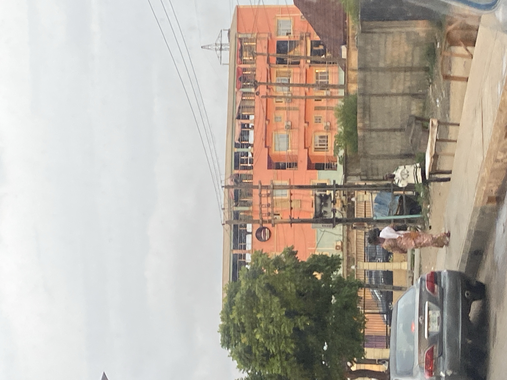A street scene in Ikeja, Nigeria, features a large, multi-story orange building with balconies and numerous windows. A mature green tree is situated to the left of the building. Power lines crisscross the sky above the scene. In the foreground, a dark gray sedan is parked on the left side of the road. A woman in a patterned dress and a white head covering walks on the sidewalk, facing away from the camera, and a man wearing a black and white striped jacket stands on the sidewalk near a wooden bench. A concrete wall runs along the right side of the scene. The sky is overcast.