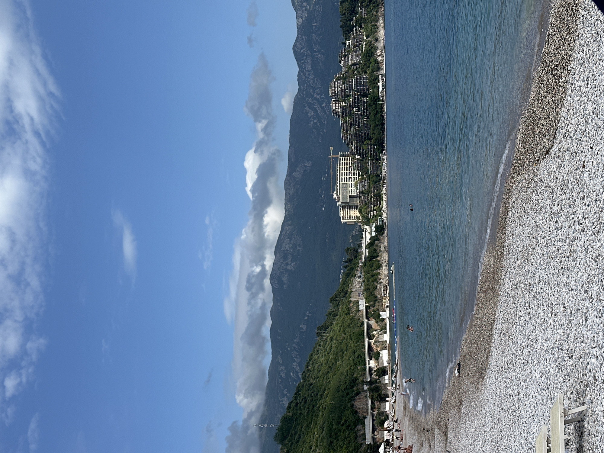 The image shows a coastal view in Budva, Montenegro. The sky is blue with scattered white clouds, and a large mountain range runs parallel to the coast, partially obscured by clouds. A dense concentration of buildings and vegetation covers the base of the mountains, extending towards the shoreline. A large, modern building with a construction crane nearby is visible. The Adriatic Sea is a clear, deep blue, with a pebble beach lining the coast. Several people are swimming and sunbathing. The beach is mostly white pebbles with some areas of darker stones near the waterline. Beach umbrellas and lounge chairs are present.