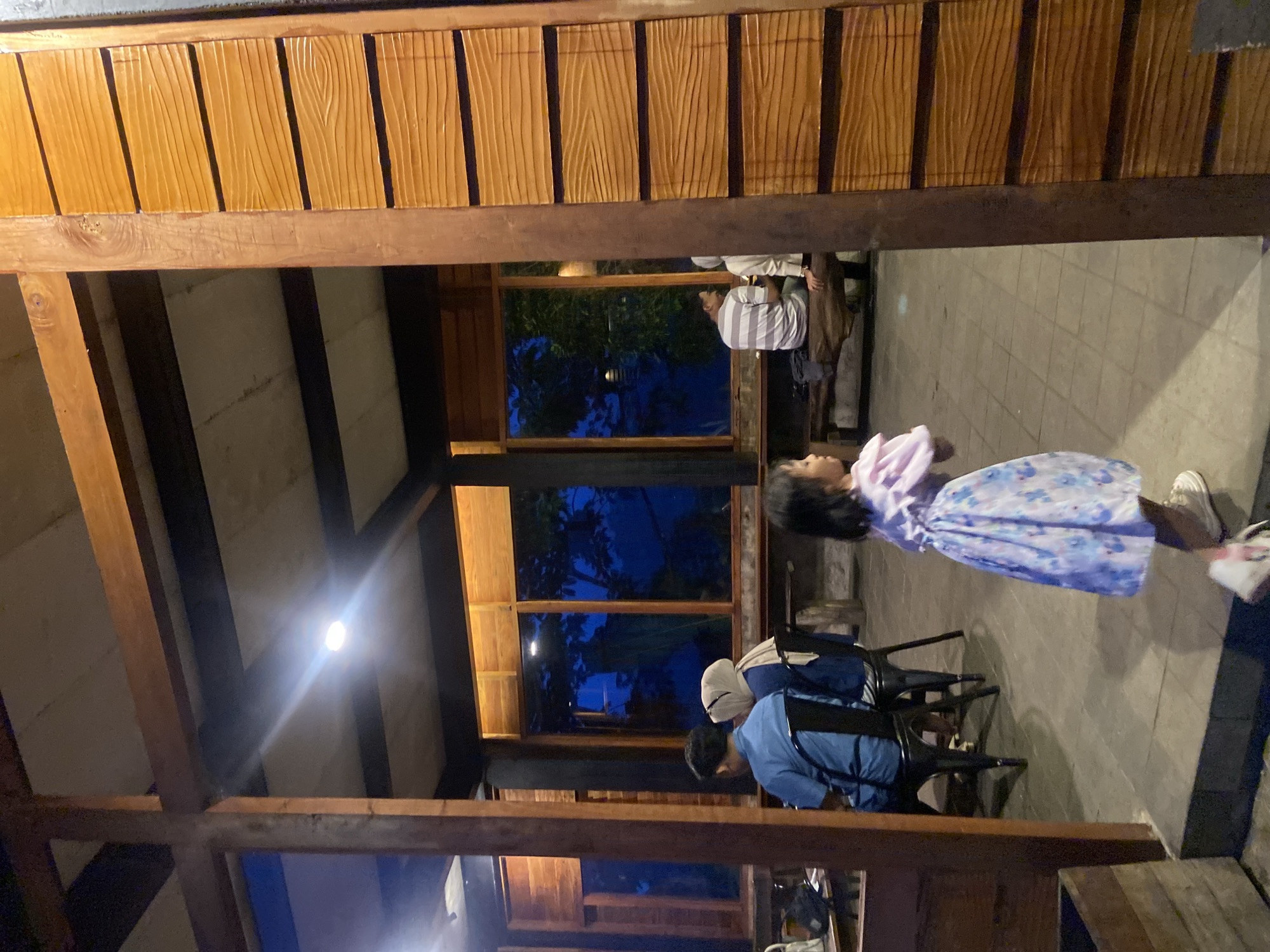 The image depicts a scene at night in what appears to be an open-air structure with a wooden frame and roof. The location may be in Tamiajeng, Indonesia. The roof is constructed with parallel wooden beams. A light fixture is visible on the ceiling.
Several people are present. A girl in a light-colored dress with a floral pattern is standing and looking toward the camera's perspective. Seated at a table are two individuals, one wearing a light blue shirt and another with a white head covering. In the background, another person in a striped shirt is seated.
The scene suggests an informal outdoor dining or gathering area. The lighting is dim. The visible environment includes outdoor vegetation seen through the structure's openings.