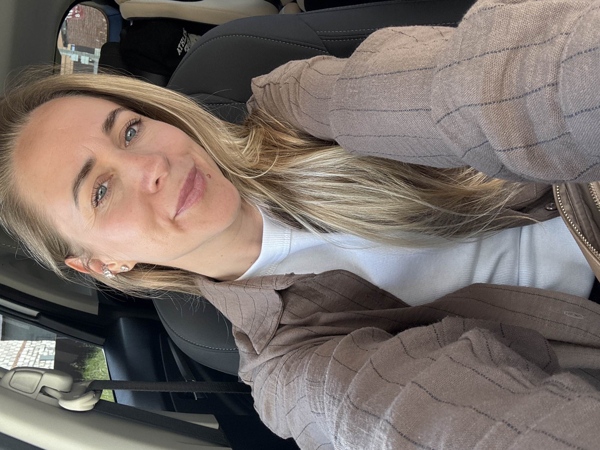 A blonde woman with blue eyes is sitting in the passenger seat of a car, facing the camera. She is wearing a brown striped button-down shirt over a white tank top and has multiple earrings in her left ear. Her hair is long and blonde, falling over her right shoulder. The interior of the car is visible, including a black leather seat and a seatbelt. Outside the car, a brick building and some greenery can be seen through the window. The woman is smiling slightly, and her head is tilted to her left. The lighting suggests it is daytime. The image was taken in Chaikovsky, Russia.