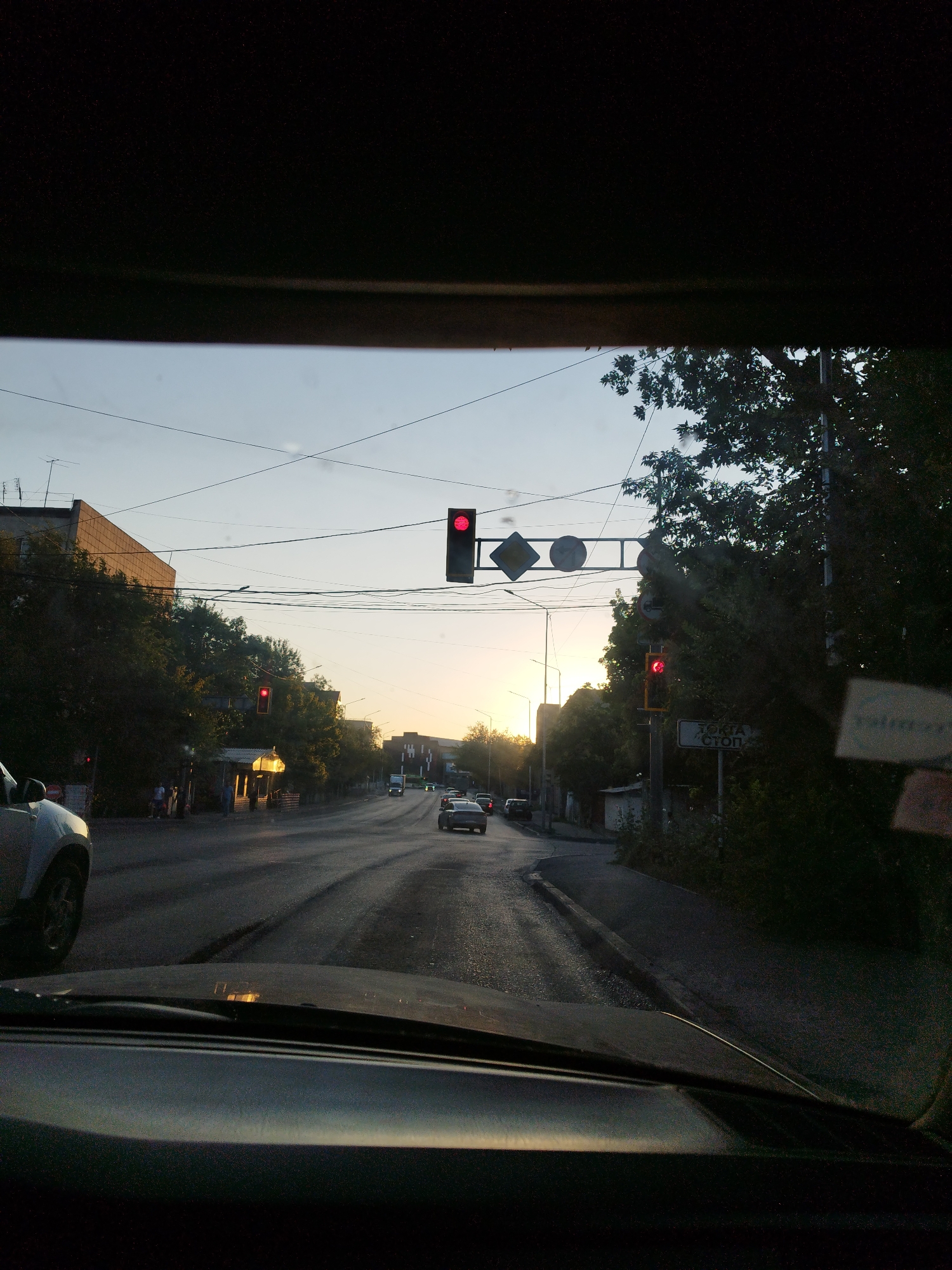 The image shows a view from inside a vehicle looking out at a street scene at dusk, likely in Shymkent, Kazakhstan. The sky is clear with a gradient of blue to orange near the horizon, indicating the sun is setting.
In the foreground, the hood and dashboard of the vehicle are visible, obscuring the lower portion of the scene. The vehicle's windshield is slightly dirty, with a few smudges or specks of dust.
The street appears to be paved and wet, with tire tracks visible. There are several cars on the road, including a silver SUV partially visible on the left, a white car further down the street, and several other vehicles further in the distance.
On the left side of the street, there are buildings and trees. One building is a multi-story structure with a textured facade. The trees are dense and leafy, casting shadows on the road. A few people are visible walking on the sidewalk near a covered area, possibly a storefront.
On the right side of the street, there are more trees and buildings. A traffic light displaying a red signal is visible hanging over the intersection. Another traffic light with a red signal is also visible on a pole further down the street. A street sign with Cyrillic lettering, reading "СТОП," is present on the right side.
Overhead, there are power lines and traffic signals suspended across the road. The overall lighting suggests it is either early morning or late evening. The scene depicts a typical urban environment with traffic, pedestrians, and infrastructure.